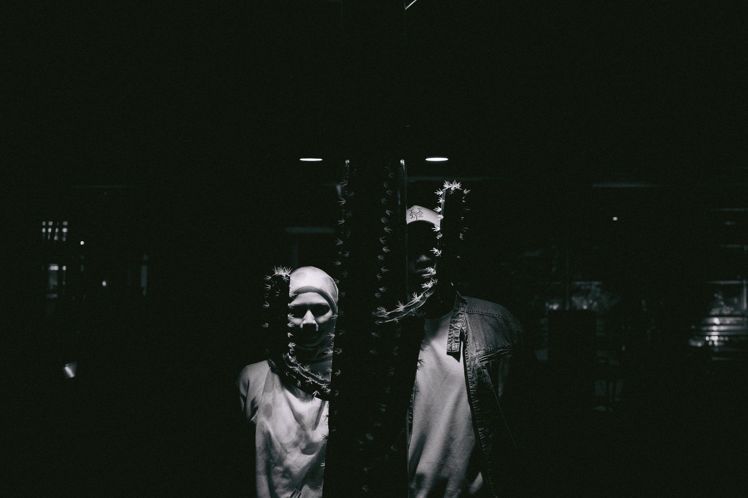 Two masked figures stand in dramatic dark lighting, creating an eerie, artistic black and white portrait.