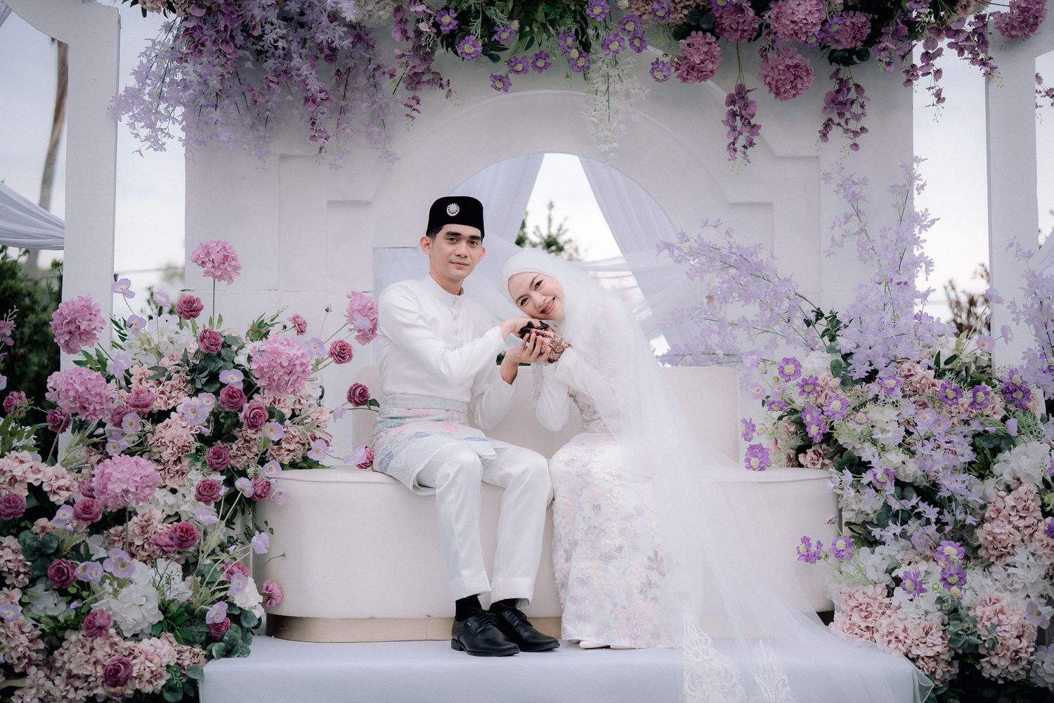 A couple in white wedding attire poses among pink and purple flowers at an elegant floral ceremony backdrop.