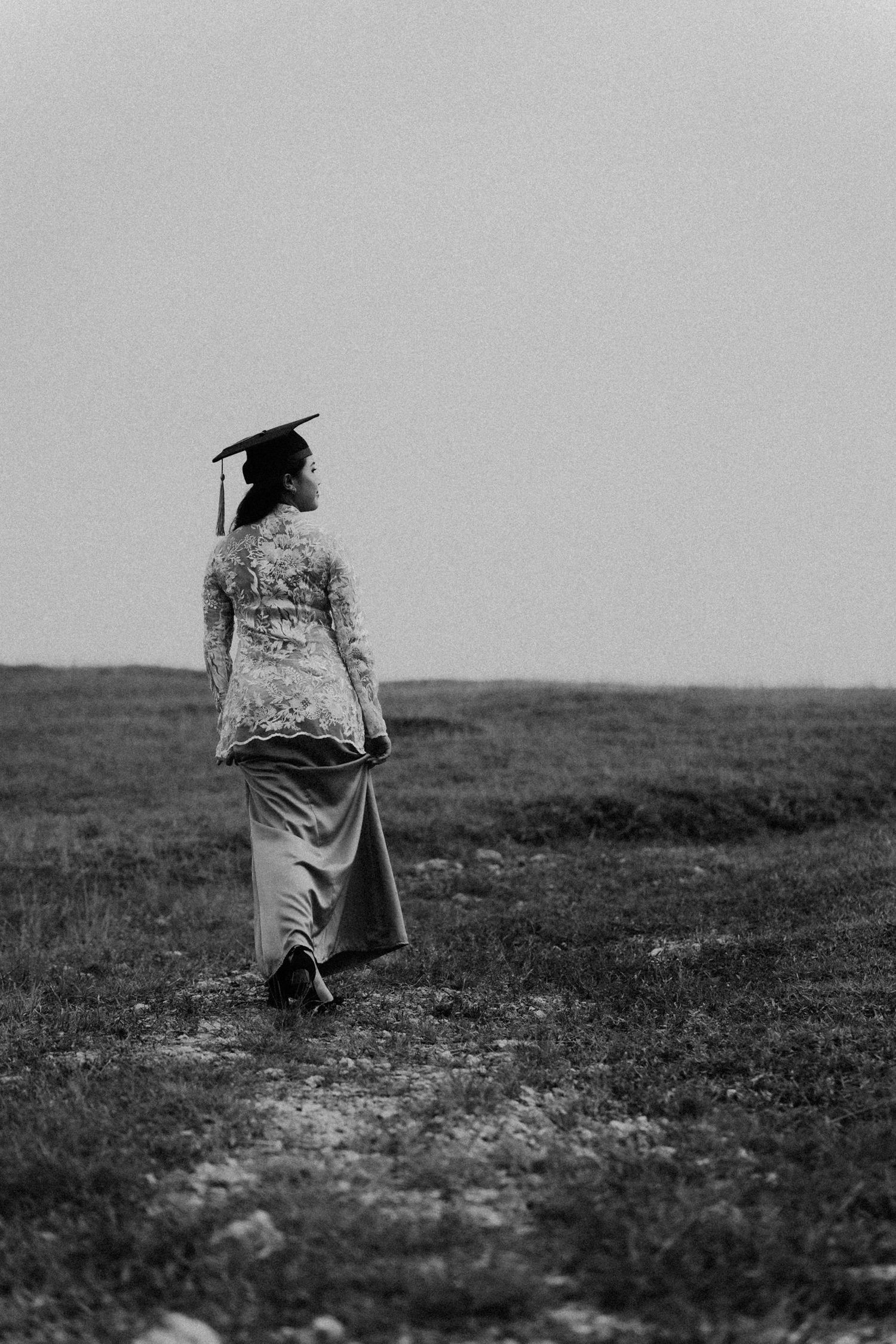 Black and white photo of graduate walking through a field wearing academic regalia and carrying diploma.