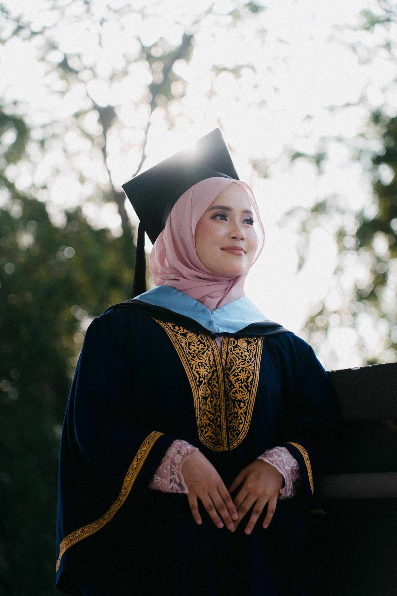 Graduate wearing black academic regalia with gold embroidery and pink hijab poses outdoors in soft natural lighting.
