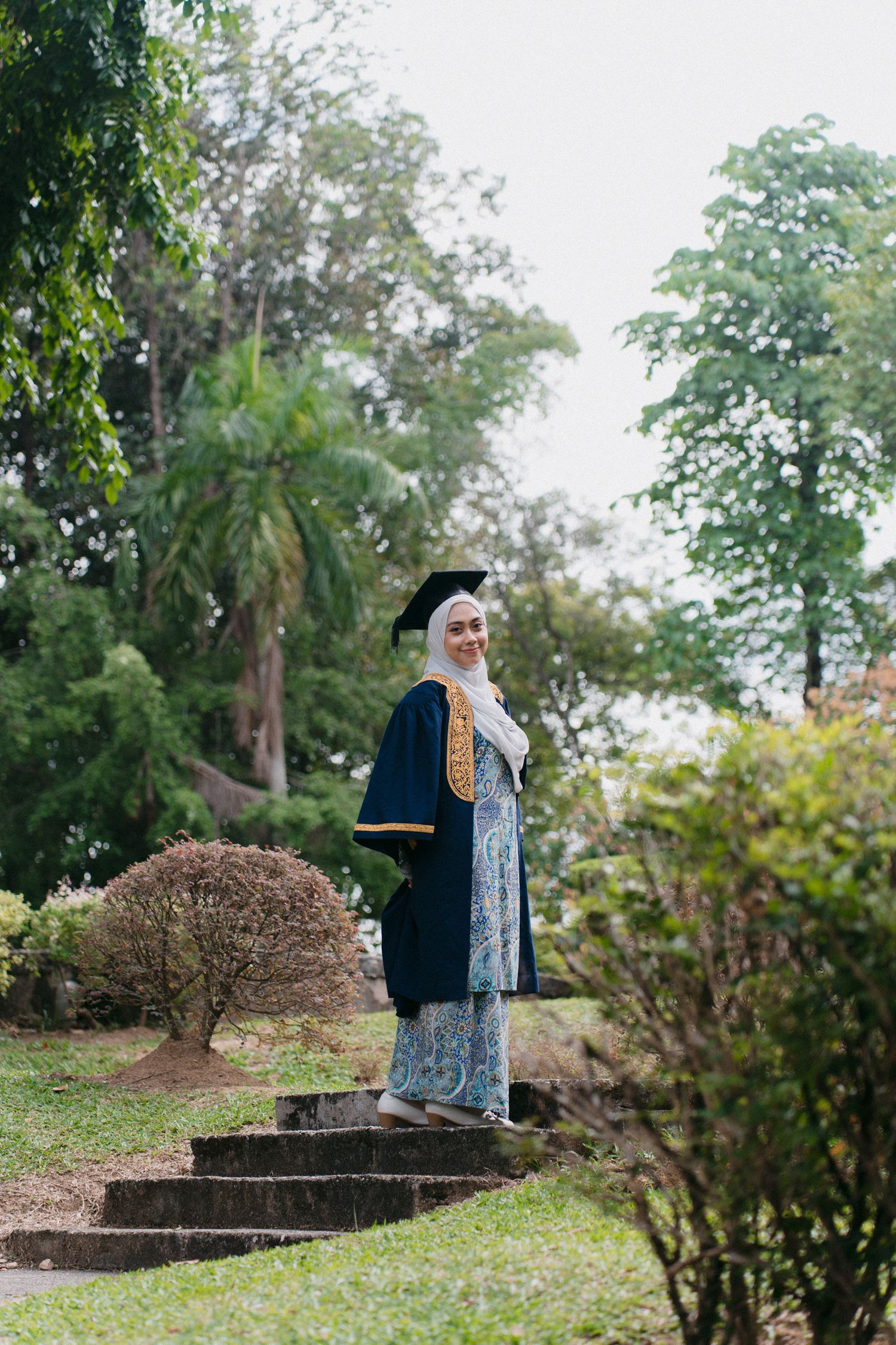 Person in blue dress and hijab standing thoughtfully in a lush garden setting with stone structures.