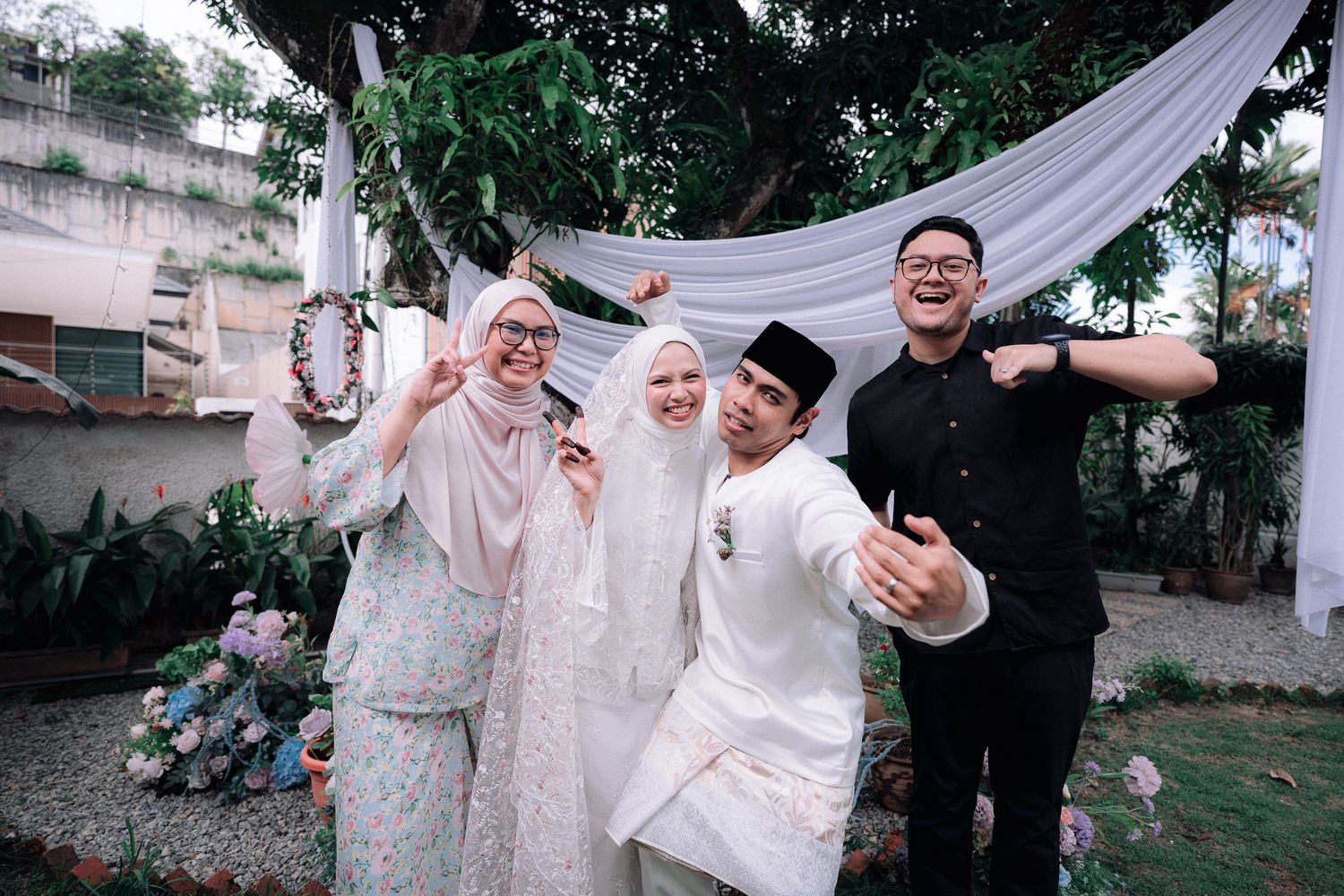 Group of four people in traditional Malay attire smiling and posing outdoors at a wedding celebration.