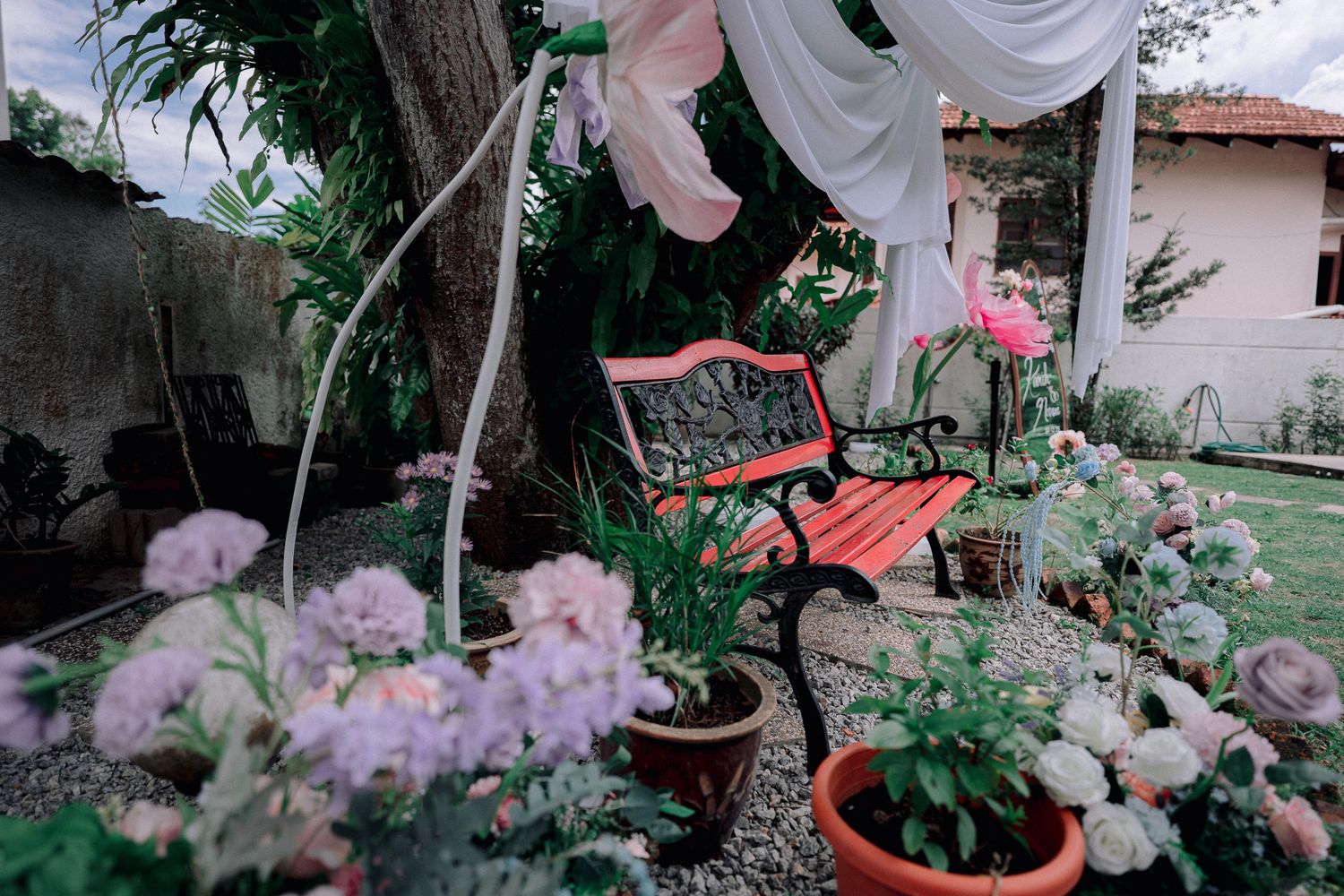 Red garden bench surrounded by blooming flowers, potted plants, and white draping fabric in a lush backyard garden.