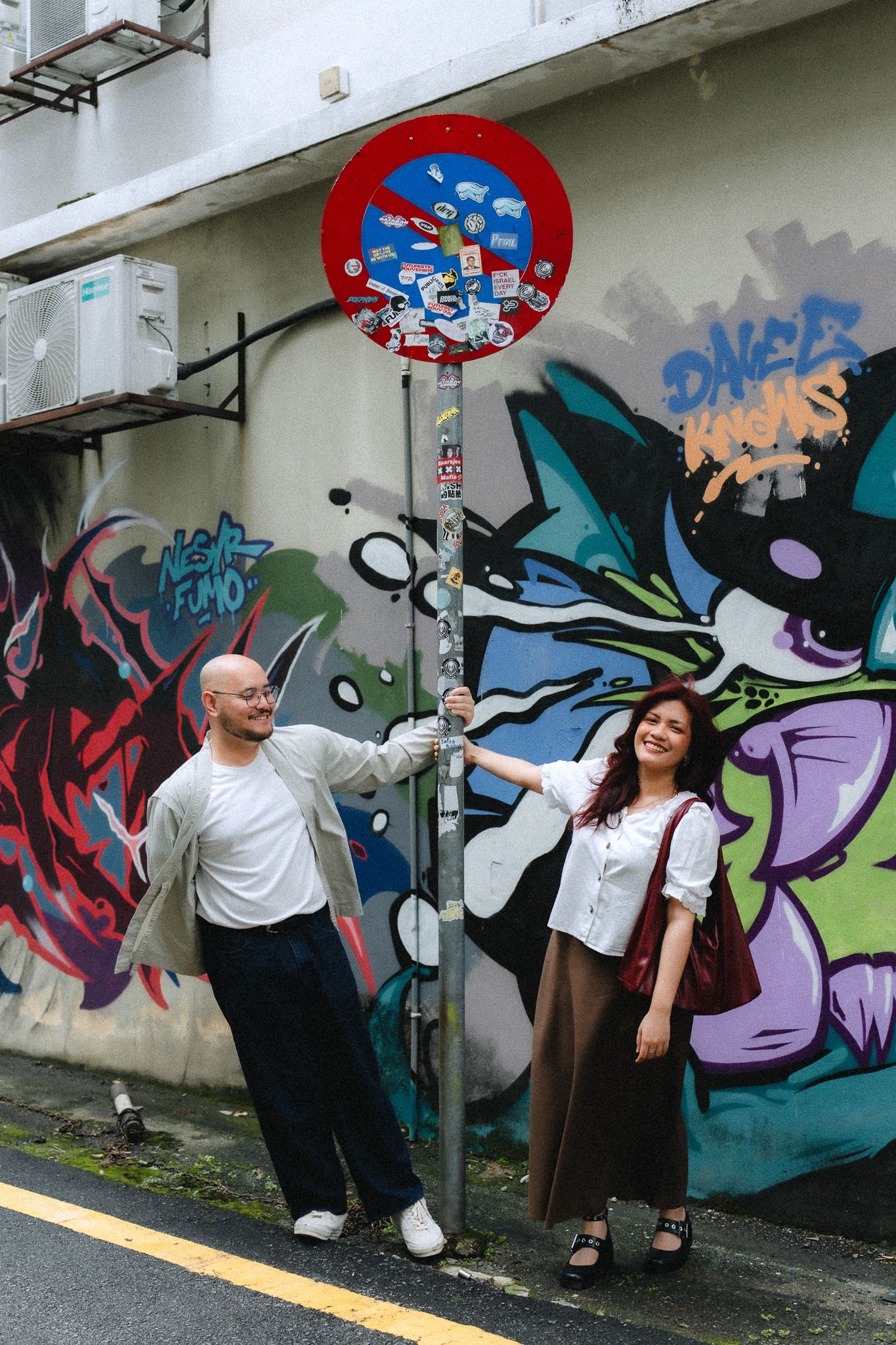 Two people pose beside a graffiti-covered street sign pole in a colorful urban alley with vibrant street art murals.