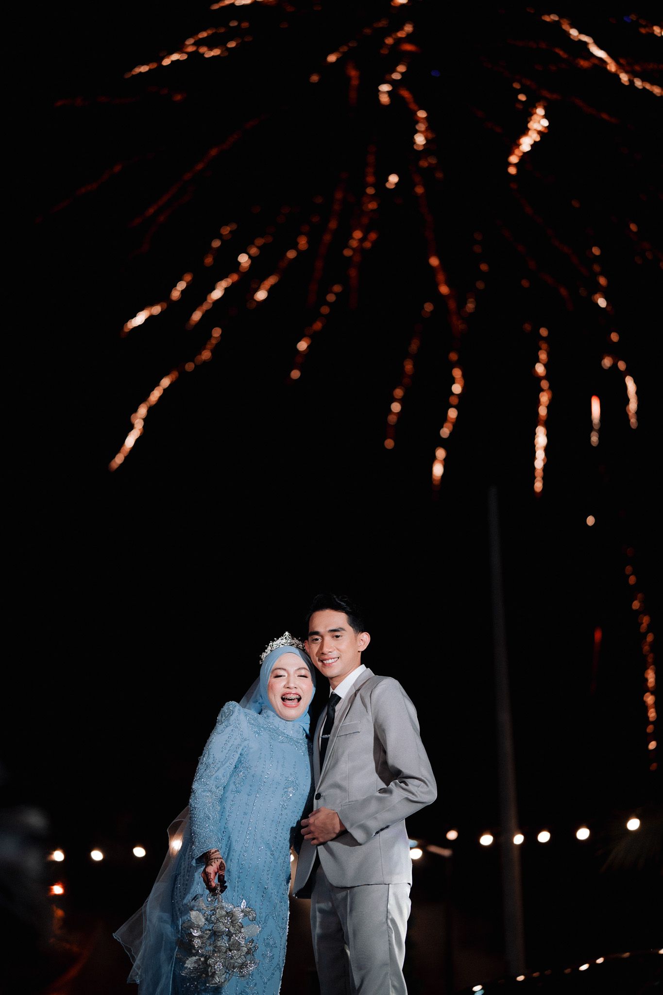 Bride in blue hijab gown and groom in gray suit pose together at night with colorful fireworks display behind them.