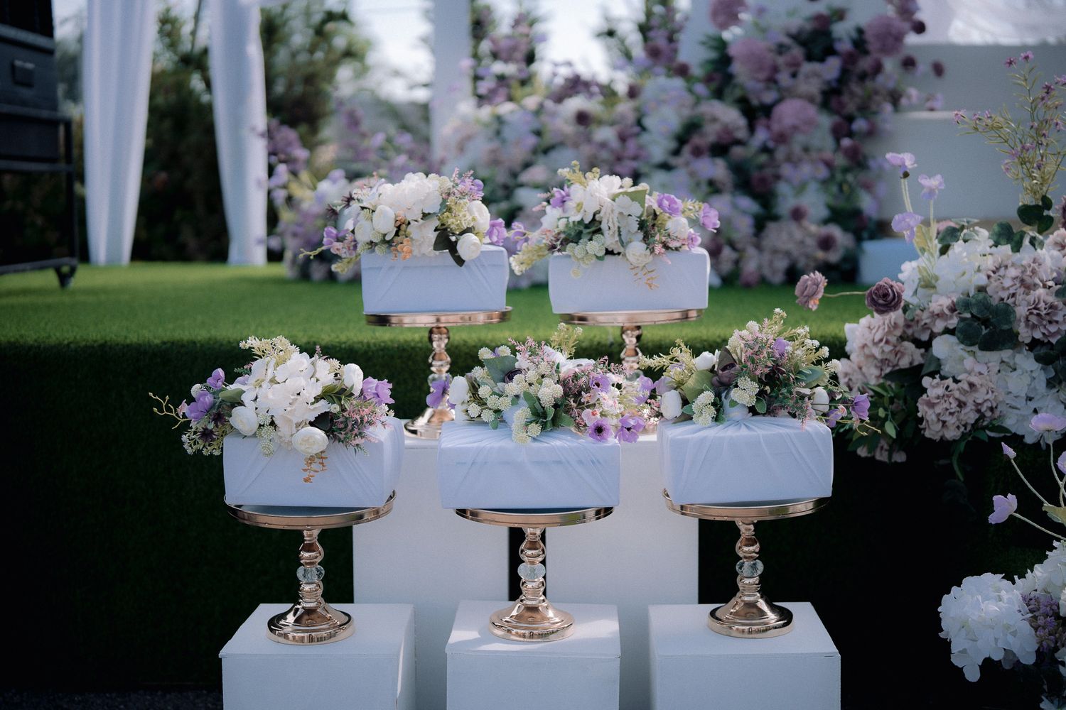 Elegant wedding cake display with white marble tiers, gold stands, and lush purple and white floral arrangements outdoors.