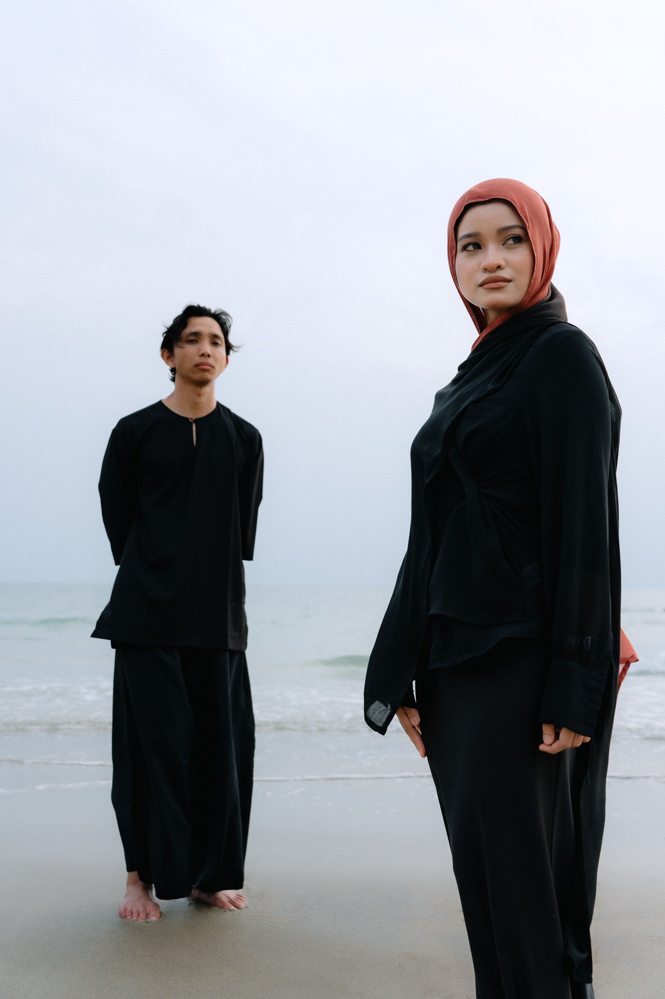 Two people in black outfits stand on a beach, woman wearing a rust hijab in foreground, man in traditional attire behind.