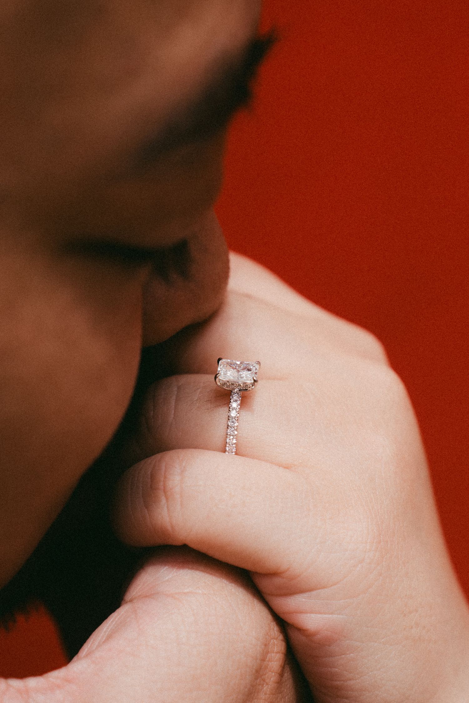 A delicate diamond engagement ring is displayed on a hand against a reddish background.