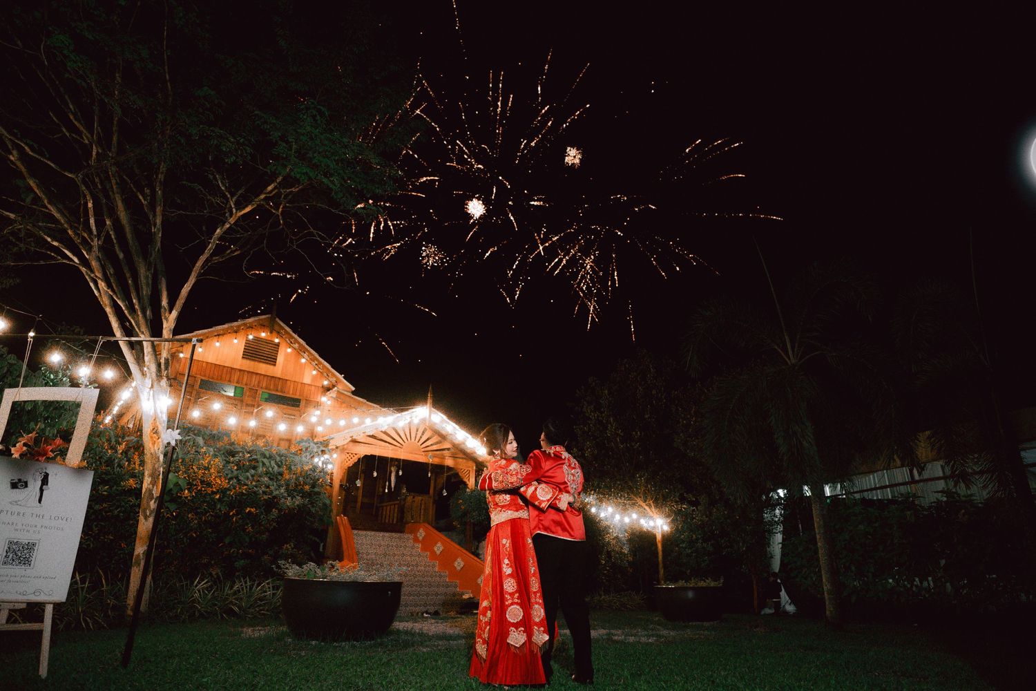 A couple in traditional Vietnamese wedding attire dance under string lights and fireworks at a backyard wedding reception.