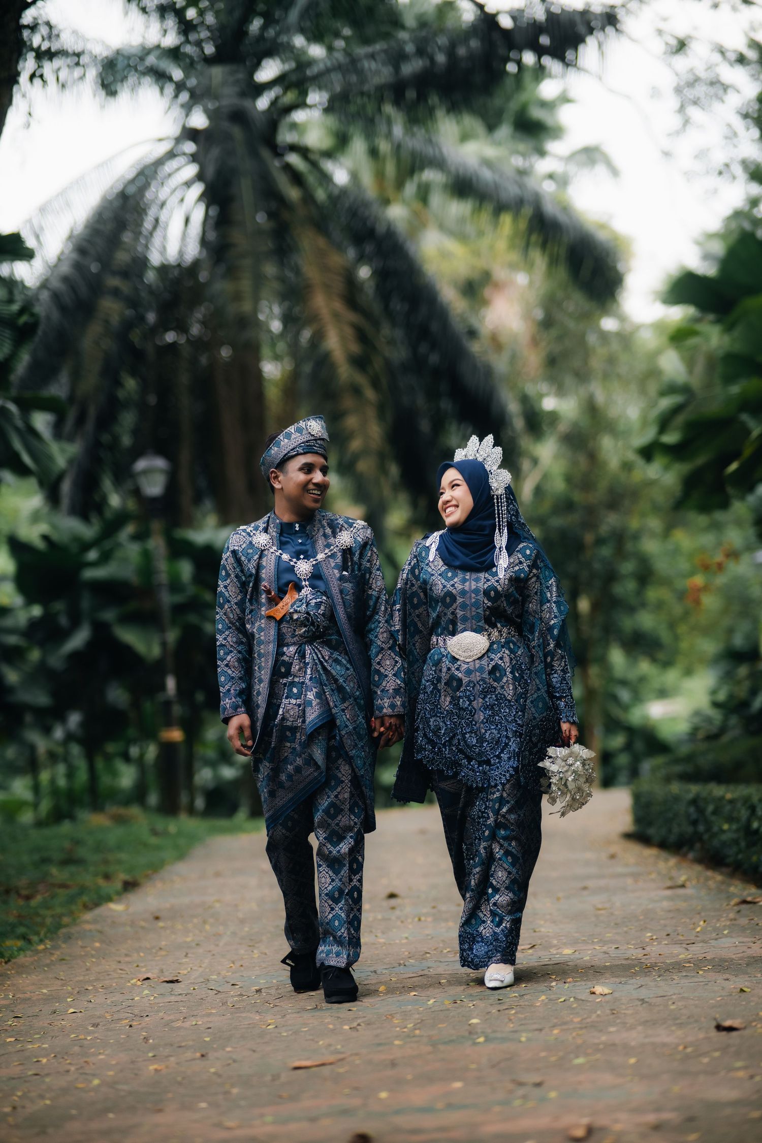 A couple wearing matching traditional blue Batik outfits walks together on a garden path surrounded by palm trees.