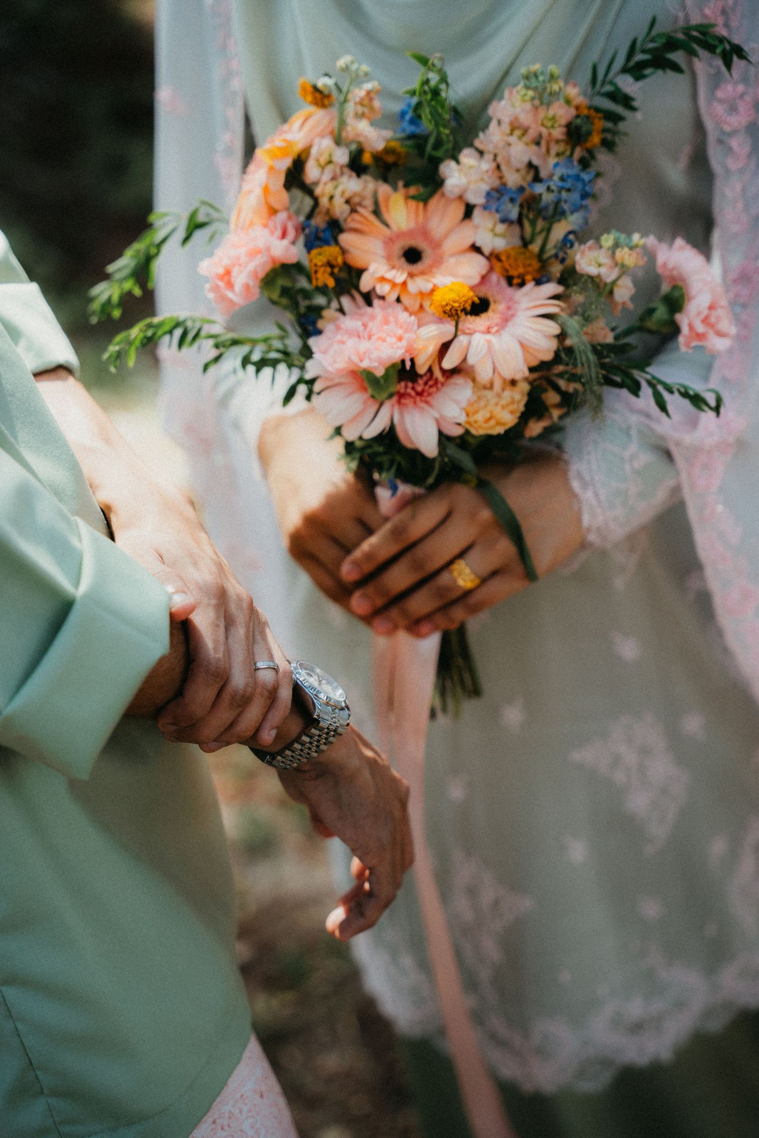 Hands holding a soft pink and peach-colored bridal bouquet with gerbera daisies and mixed flowers against lace fabric.