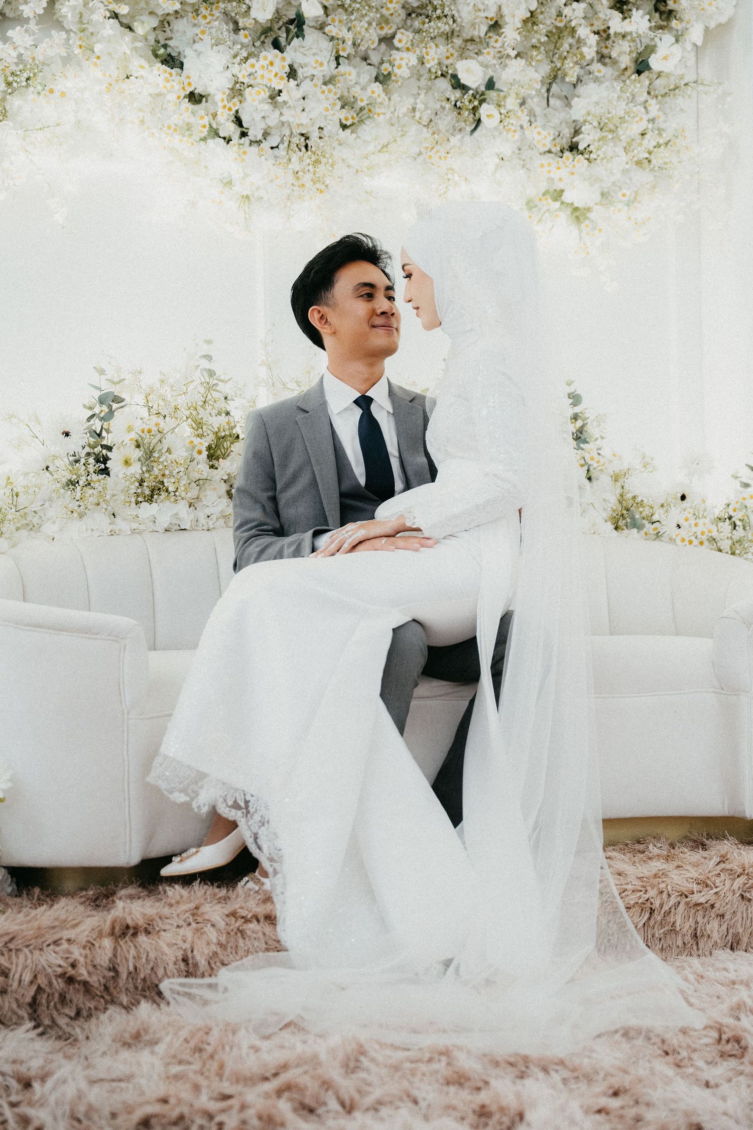 A person in a gray suit sits elegantly on a white couch surrounded by floral decor at a wedding venue.
