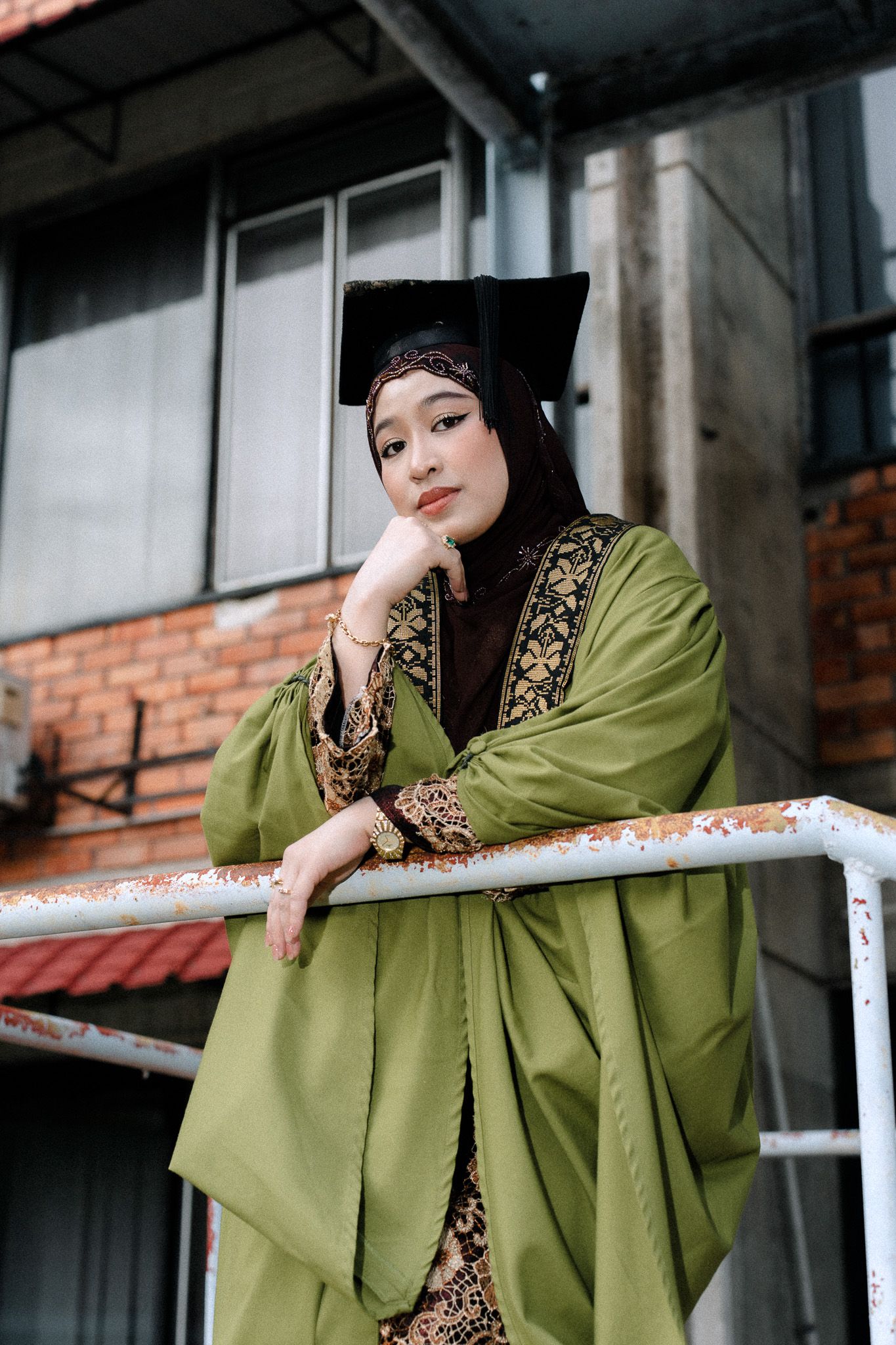 A young Muslim woman in green graduation robe and black hijab cap poses thoughtfully against a railing outdoors.