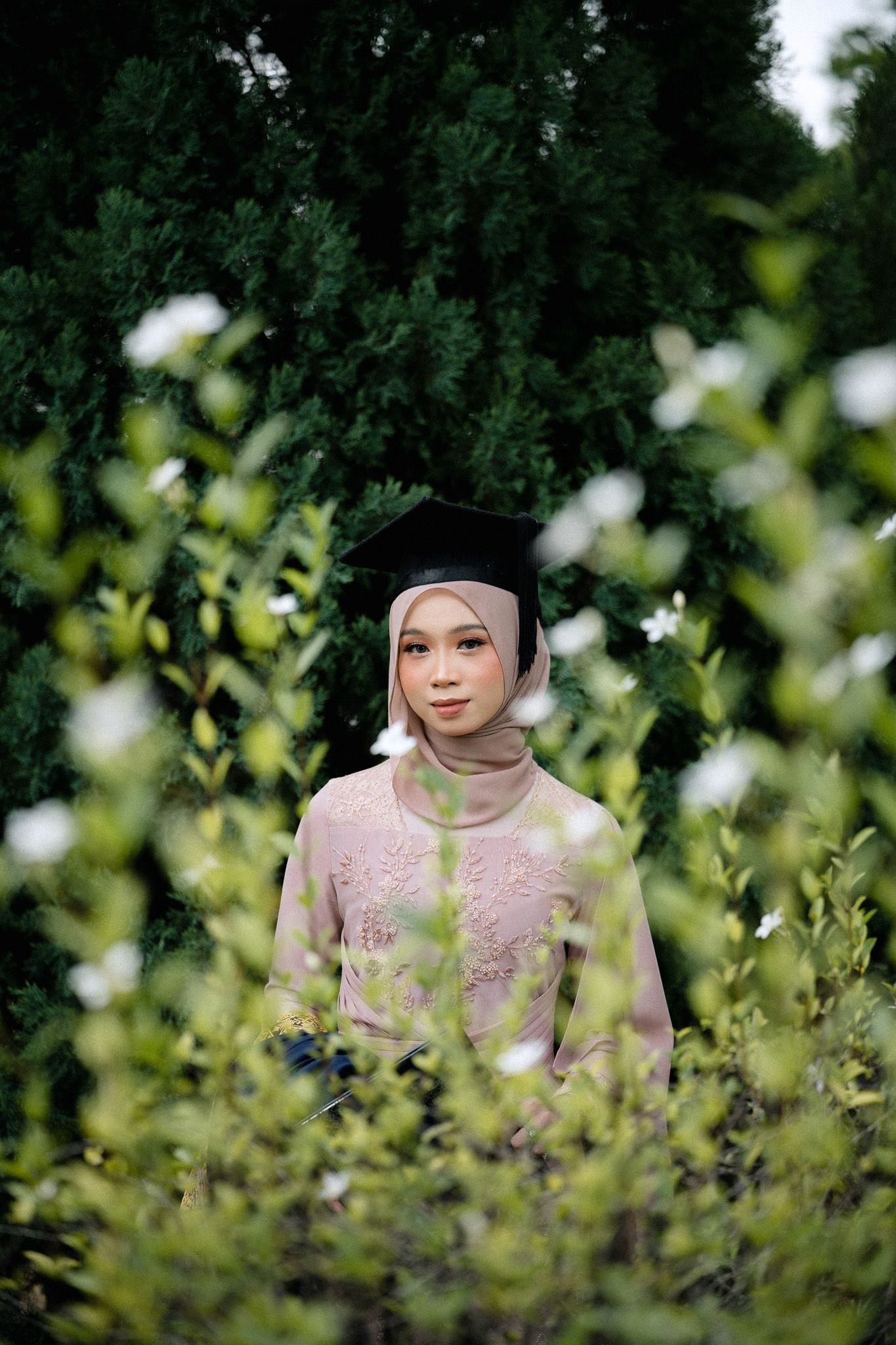 Muslim woman in hijab and graduation cap poses among lush green bushes and white flowers in pink kebaya dress.