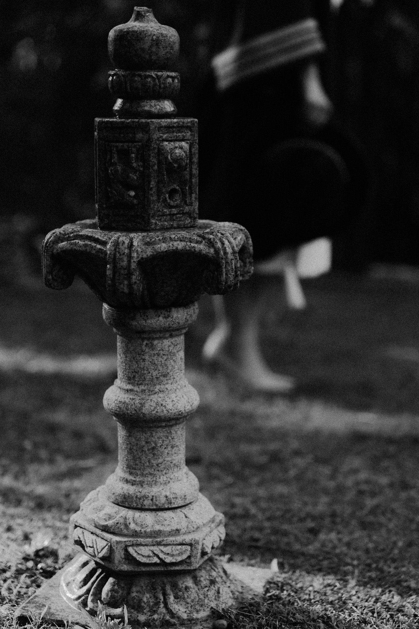 Black and white photo of an ornate stone Japanese garden lantern with blurred figures in the background.
