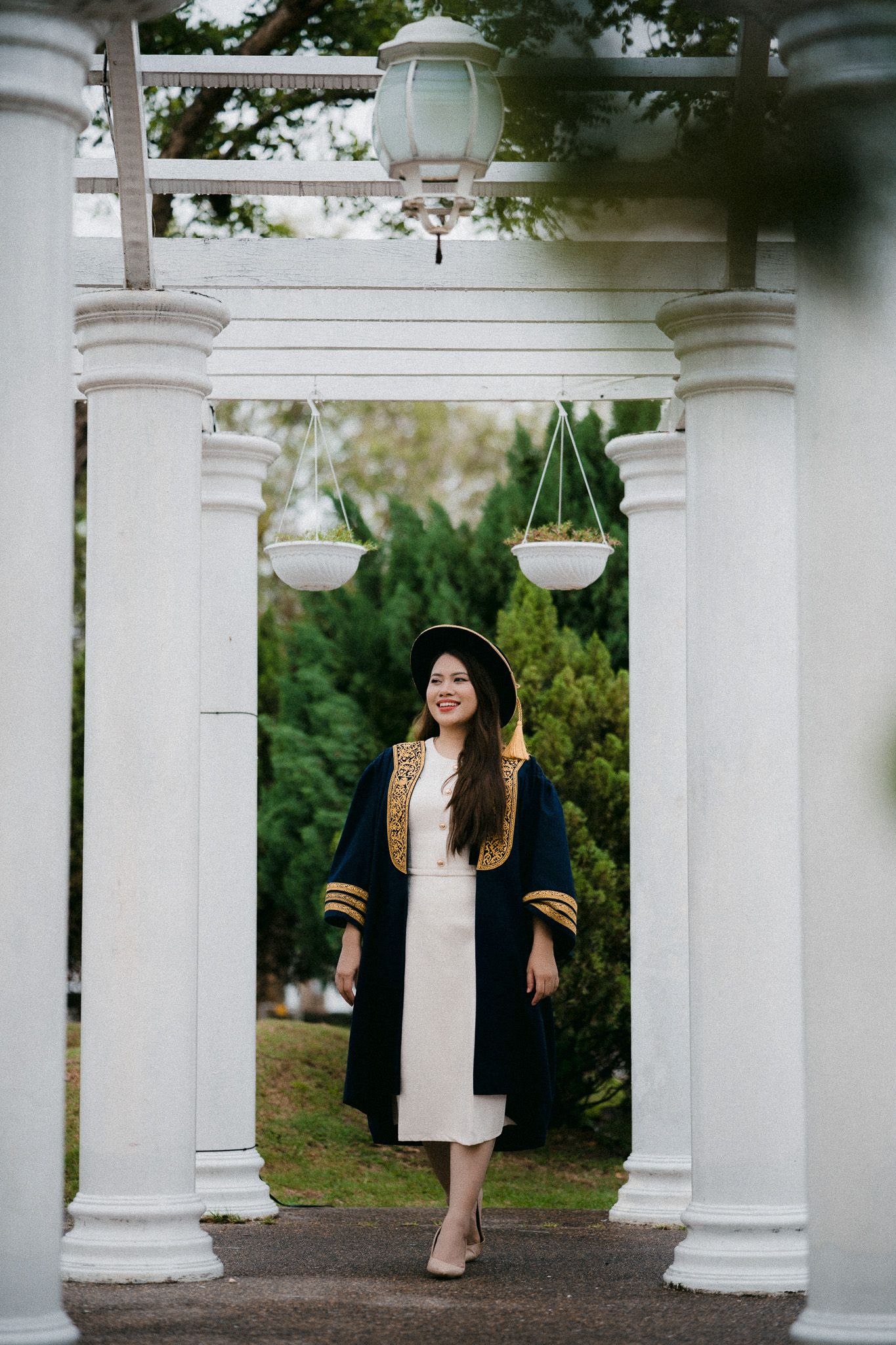 A smiling graduate in black and gold academic gown and mortarboard poses between white columns in a garden setting.
