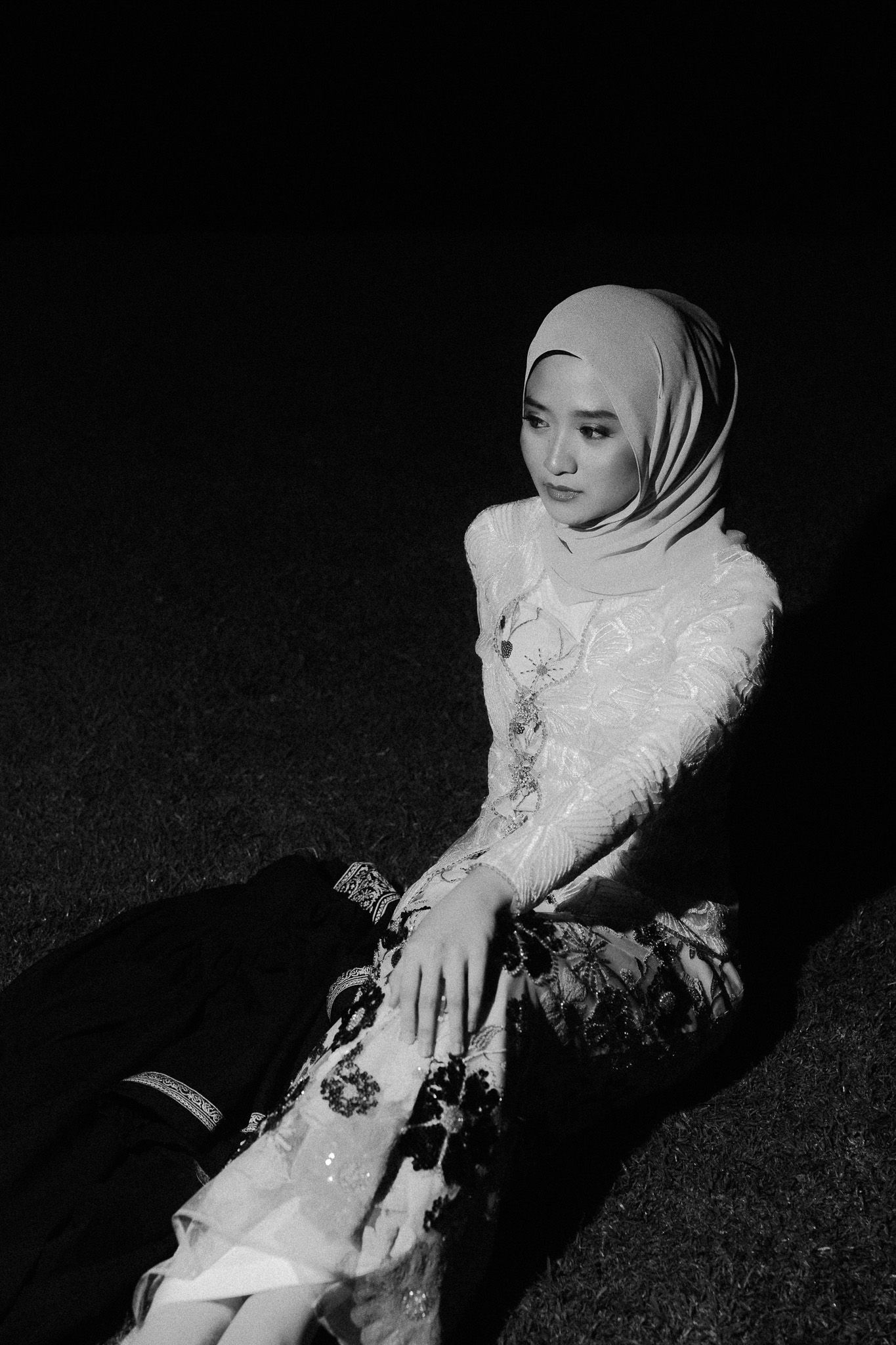 Black and white portrait of a young woman in hijab and floral dress sitting gracefully on the ground at night.