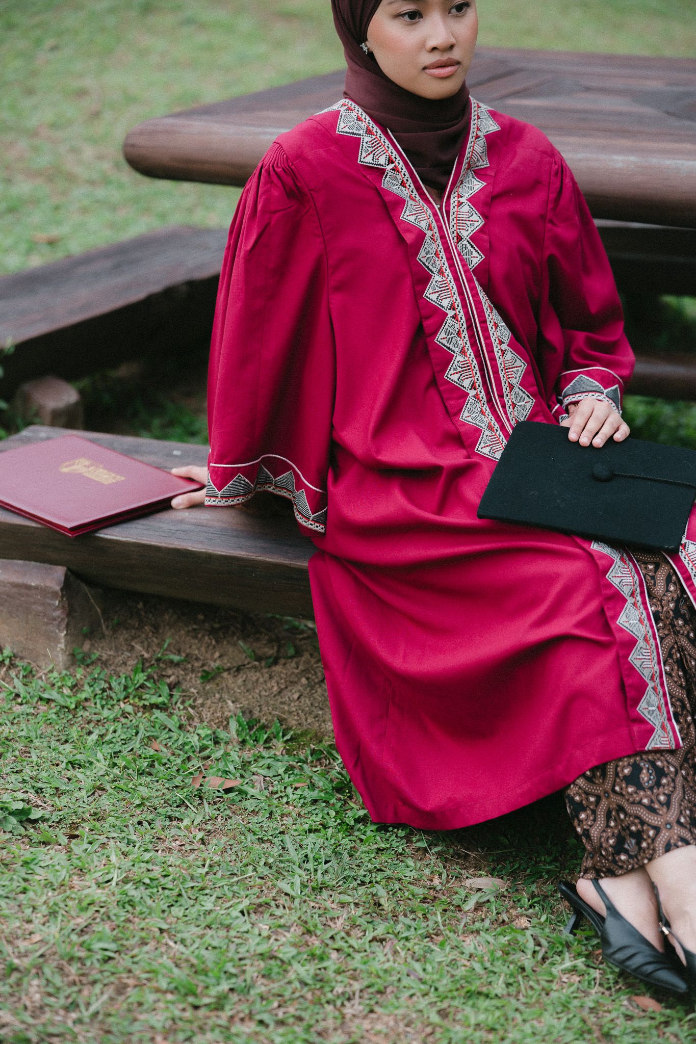 Young Muslim woman in red embroidered dress holding diploma and black folder, sitting on wooden bench outdoors.