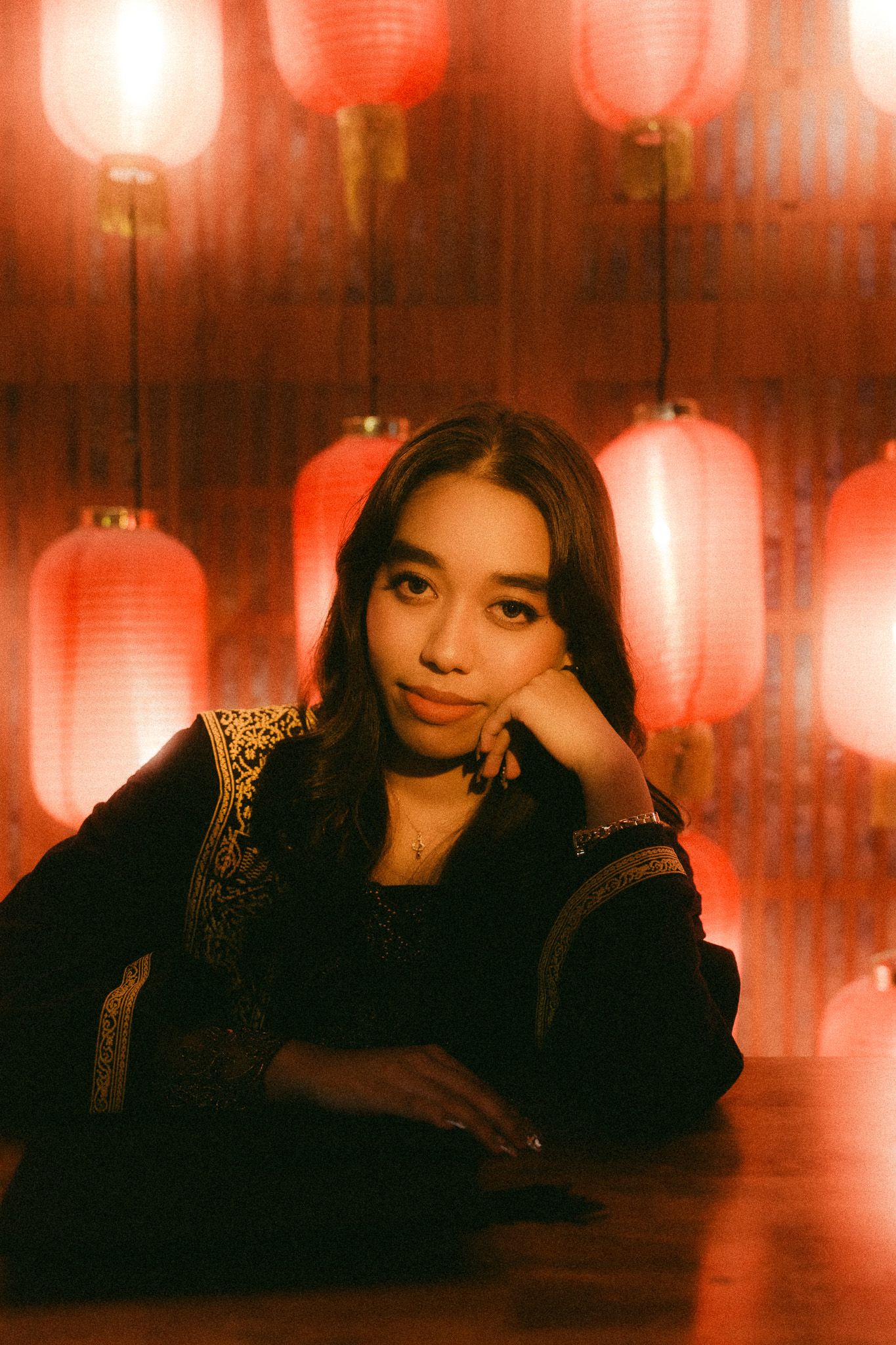 Young woman in black dress poses thoughtfully against glowing red Chinese lanterns backdrop in warm ambient lighting.