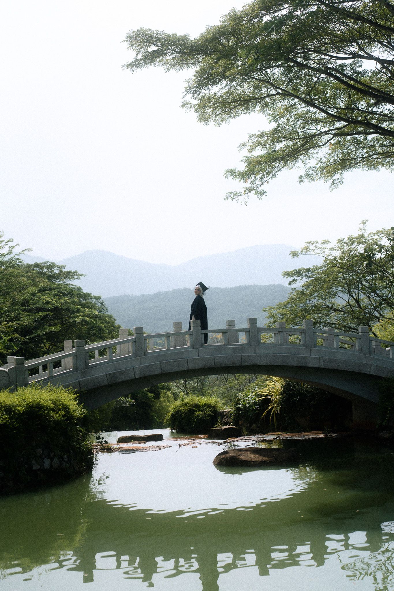 Person in traditional attire standing on a stone arch bridge over a serene green pond surrounded by lush trees and mountains.