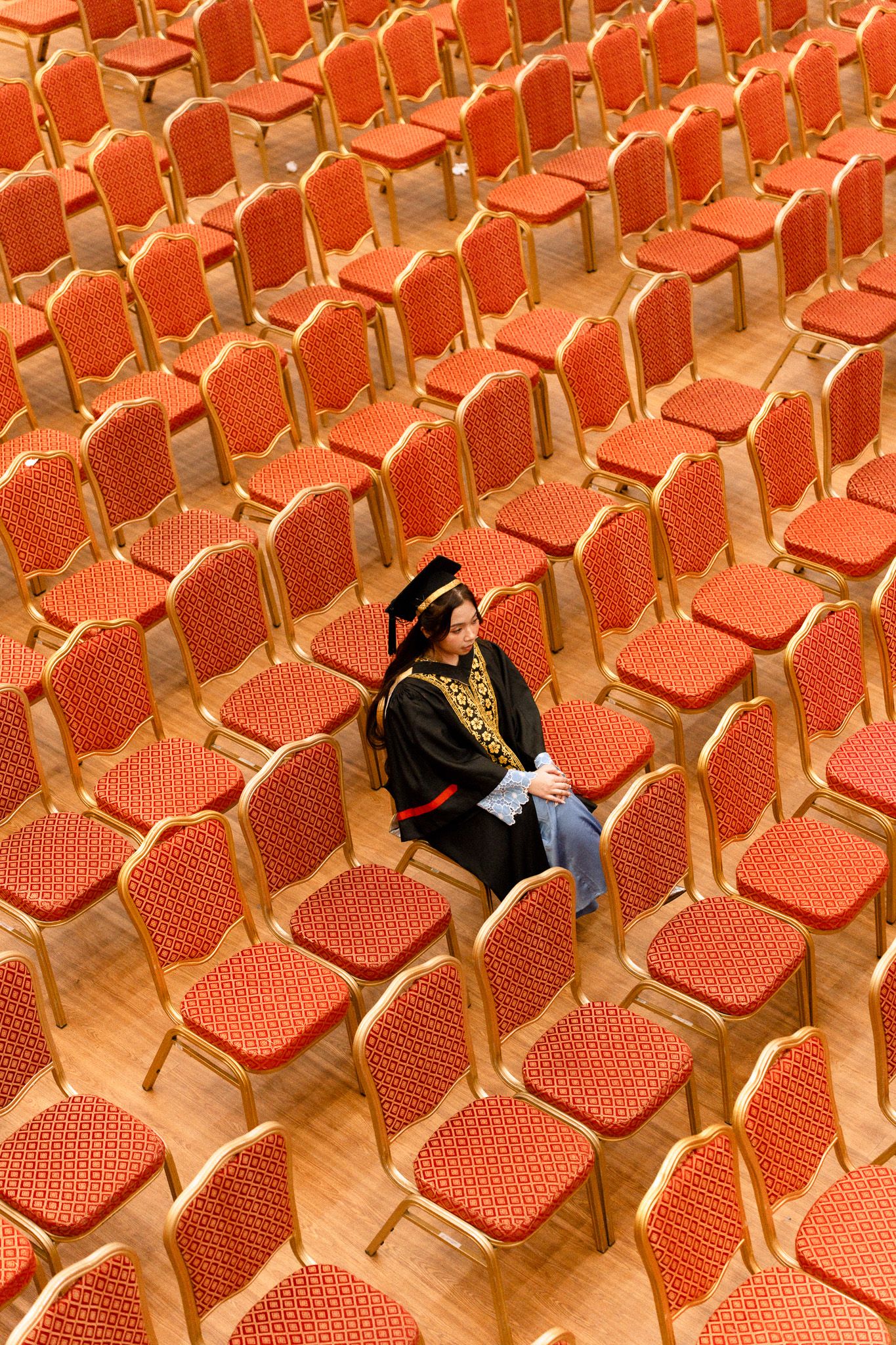 Graduate in academic regalia sits alone among rows of empty red chairs in a large ceremonial hall.
