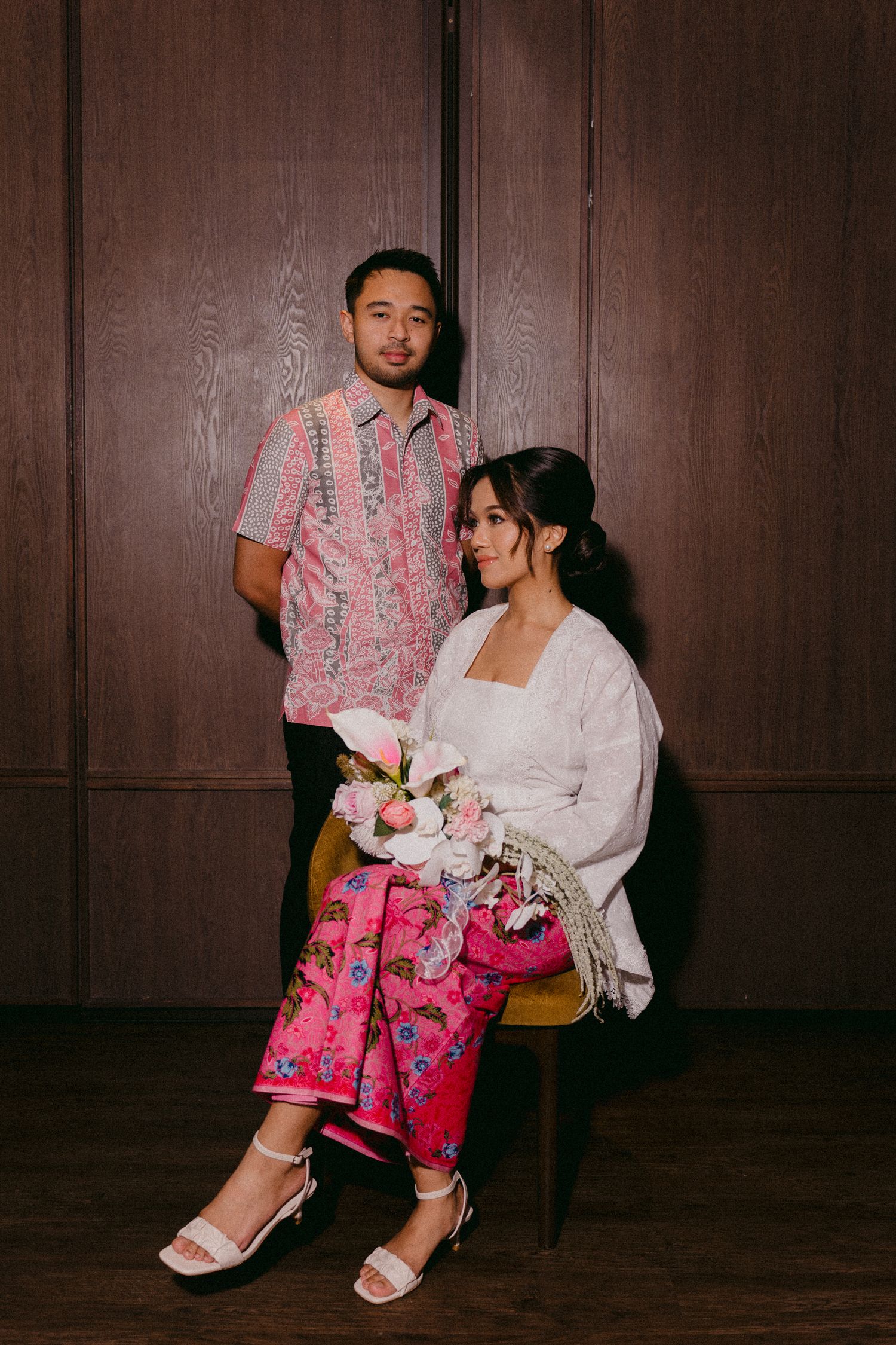 A couple poses against wood paneling wearing pink floral patterns, with traditional formal attire and white sandals.