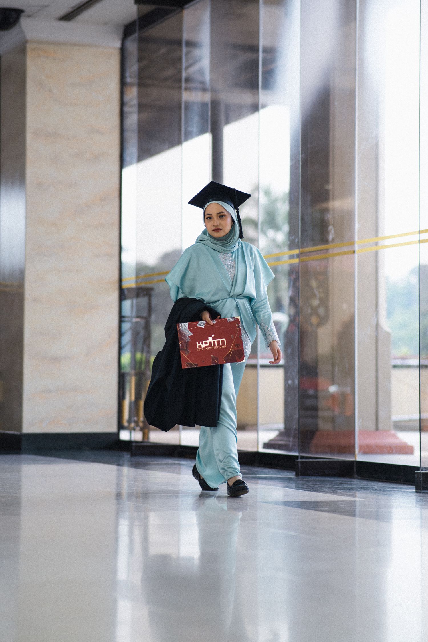 A graduate in turquoise hijab and gown walks through a modern building hallway carrying a red folder.
