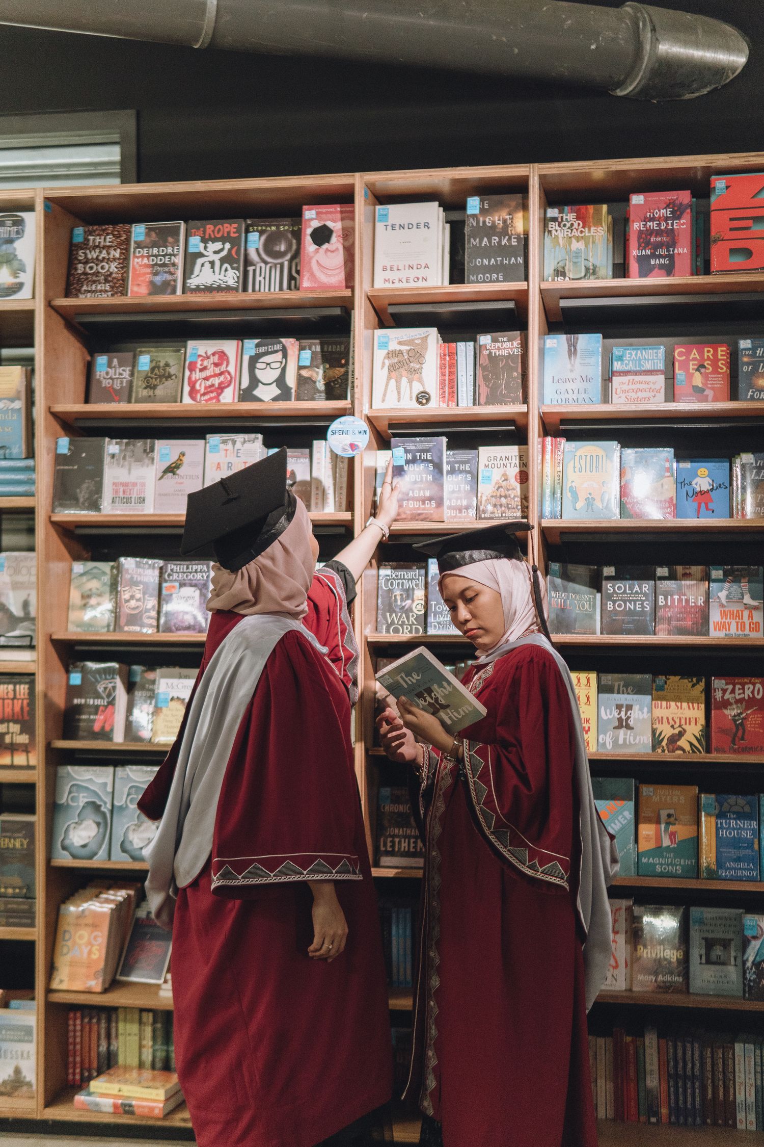 Two graduates in red robes browse books together in a library.
