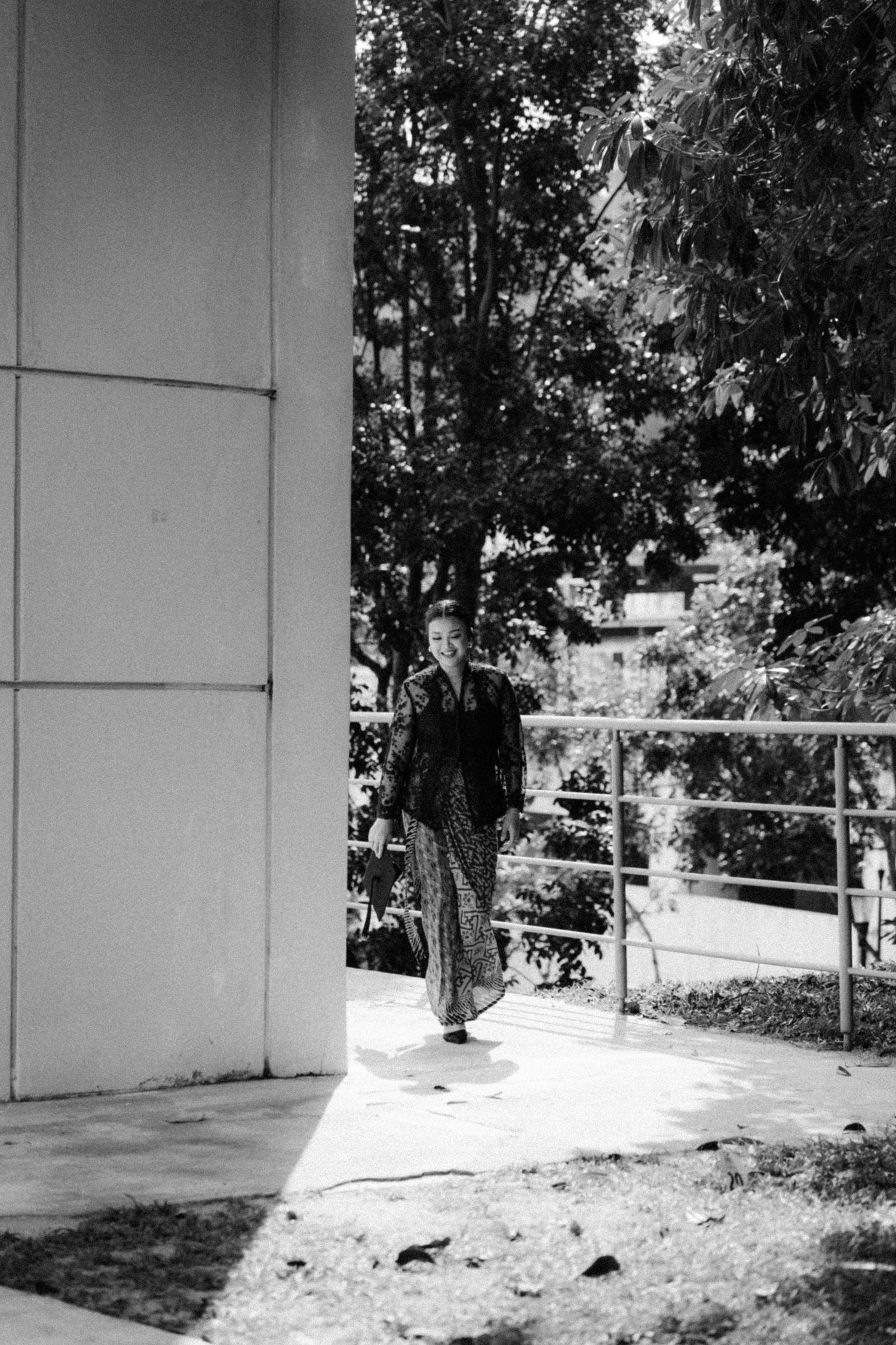 A person stands near a white pillar on a concrete walkway with trees and railing in the background in black and white.