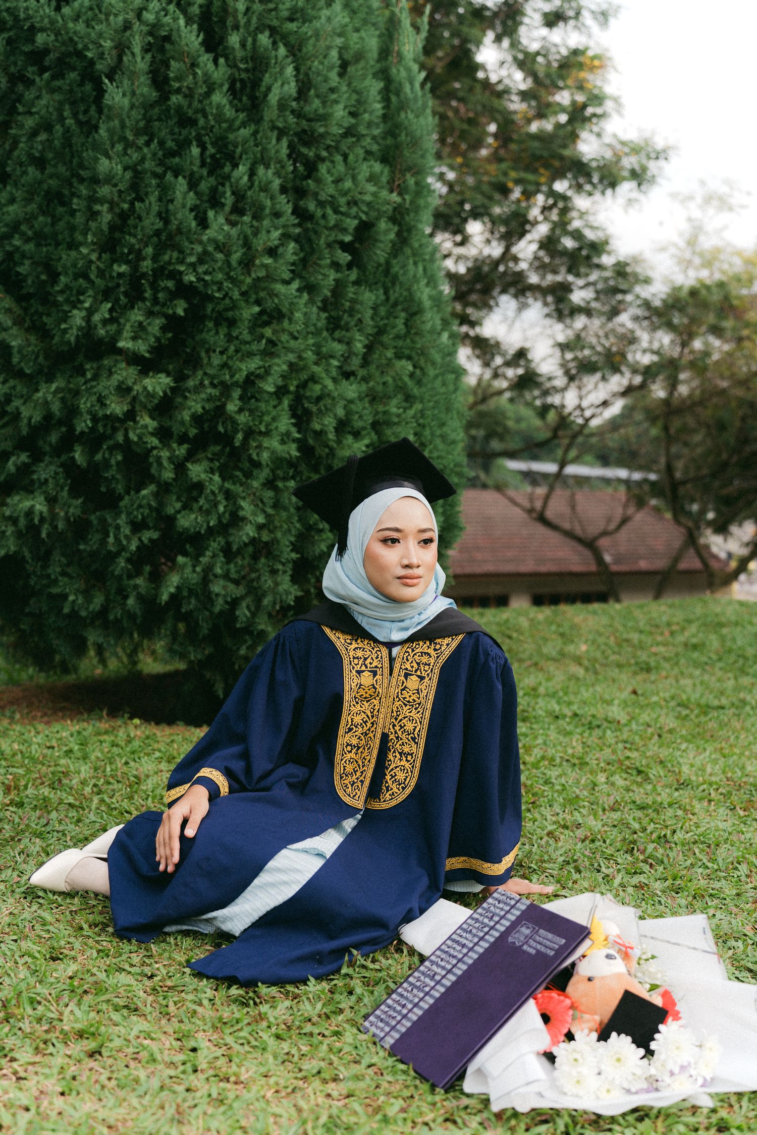 Graduate wearing hijab and navy graduation gown with gold embroidery sits on lawn with notebooks and decorations.