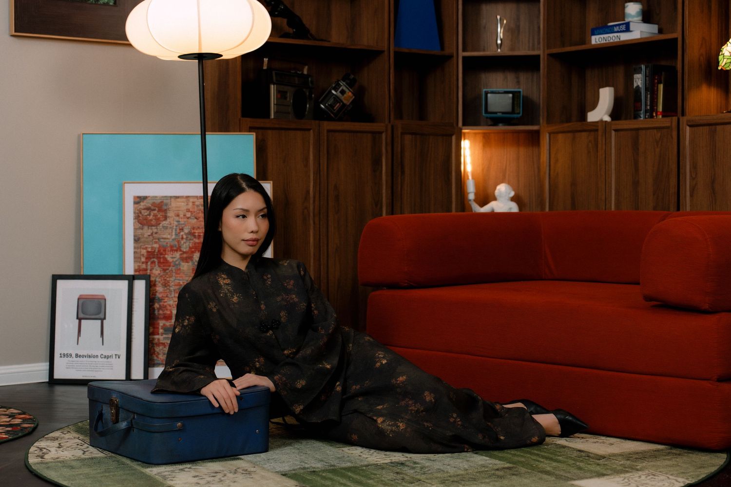 A person in a dark dress sits on a green rug next to a red couch in a wood-paneled room with vintage decor.