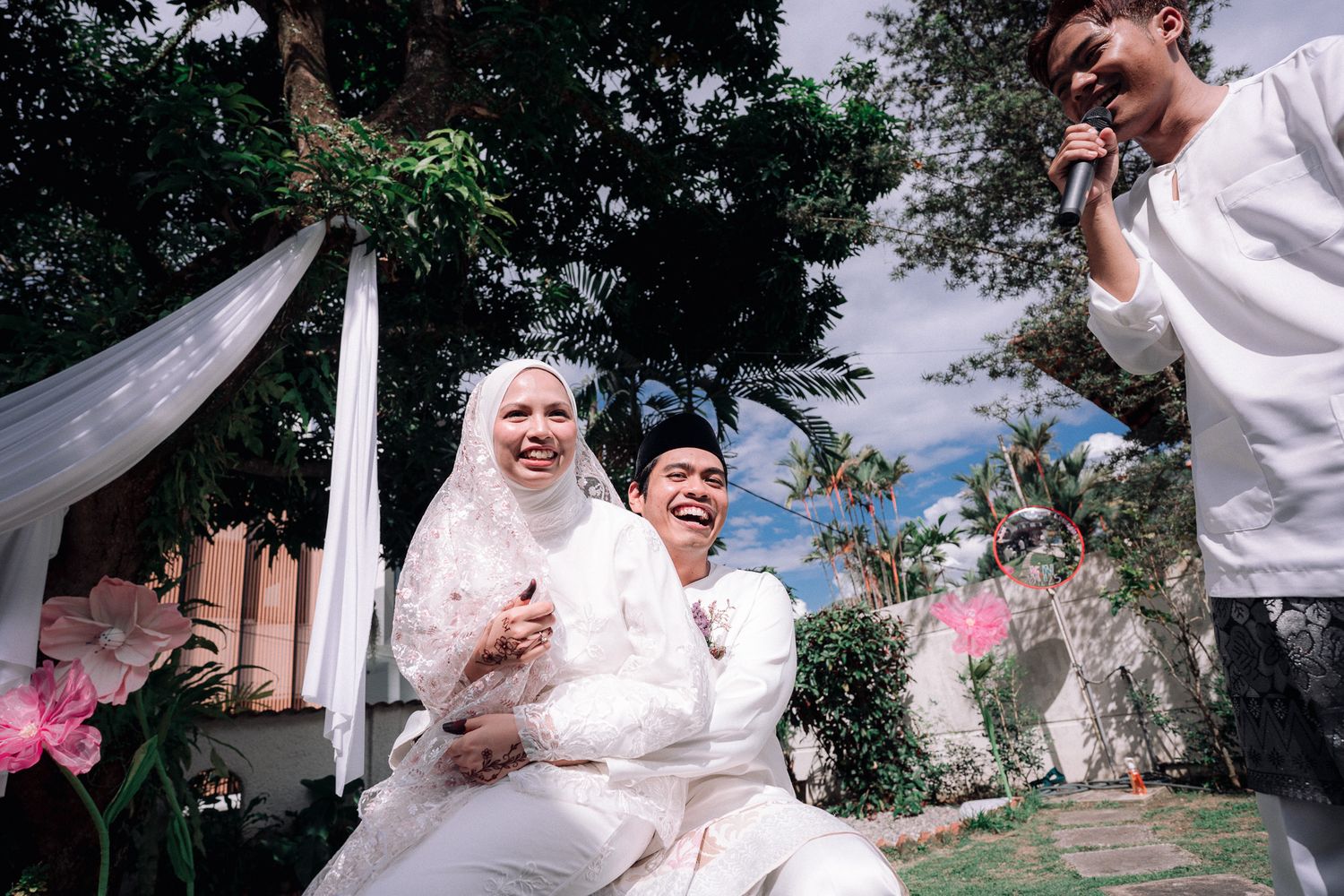 A joyful couple in white traditional wedding attire laughing outdoors at a garden ceremony.