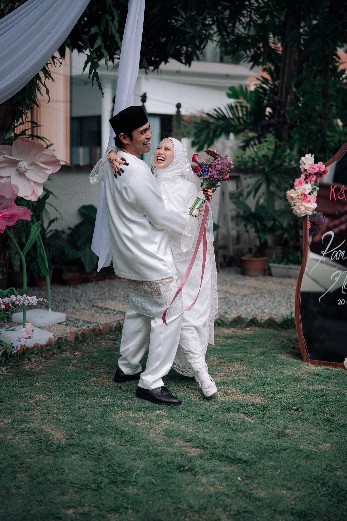 Muslim couple in white traditional attire embracing joyfully at an outdoor wedding ceremony surrounded by floral decorations.