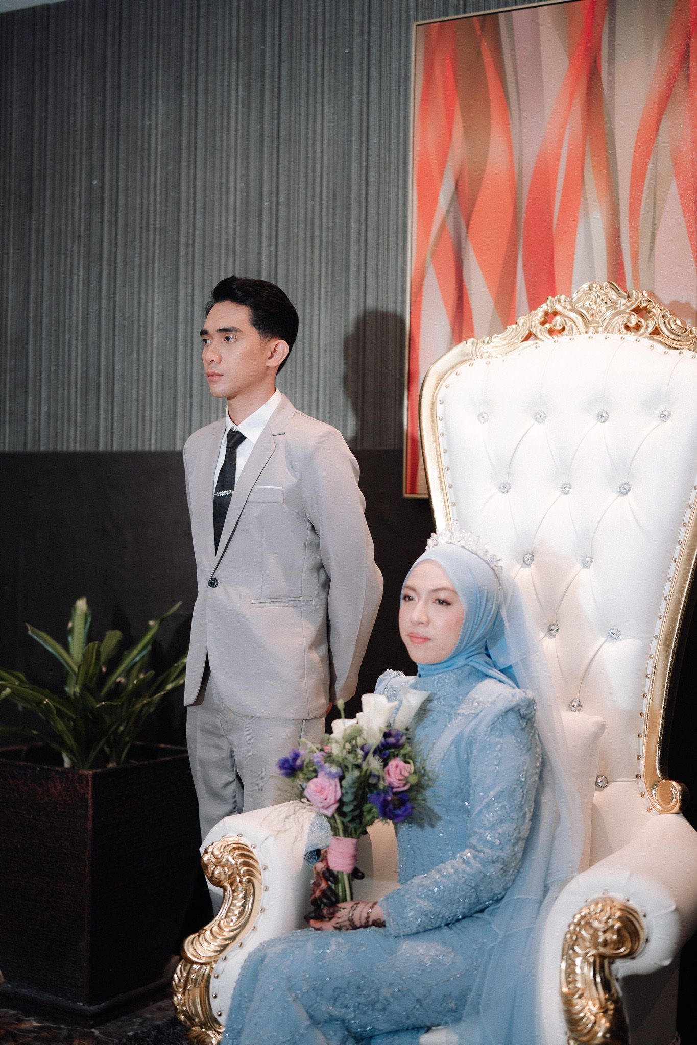 Muslim bride in blue hijab and gown holds flowers seated on ornate white throne chair beside groom in gray suit.