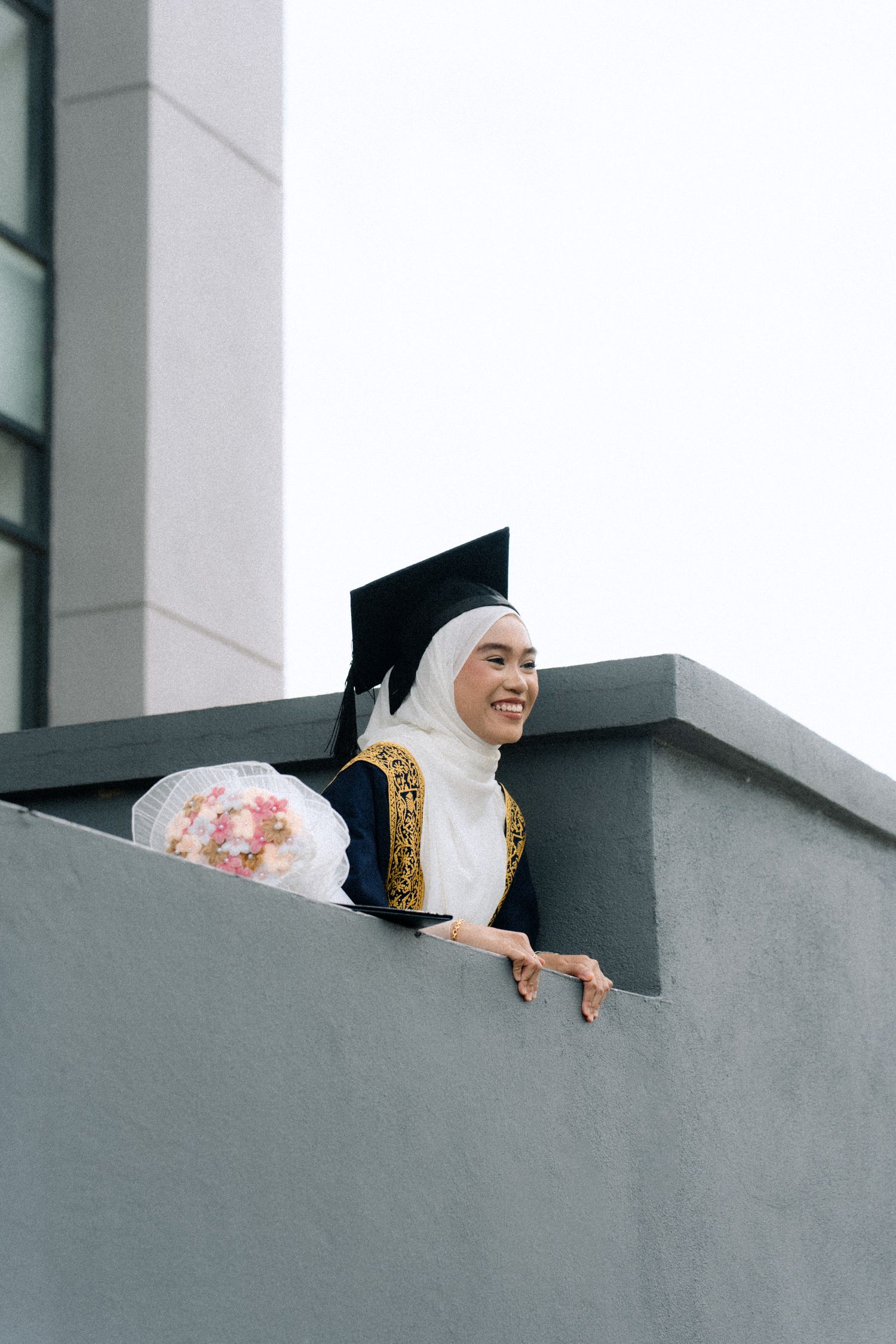 Graduate in traditional academic dress and hijab creates fun poses on a modern architectural balcony.