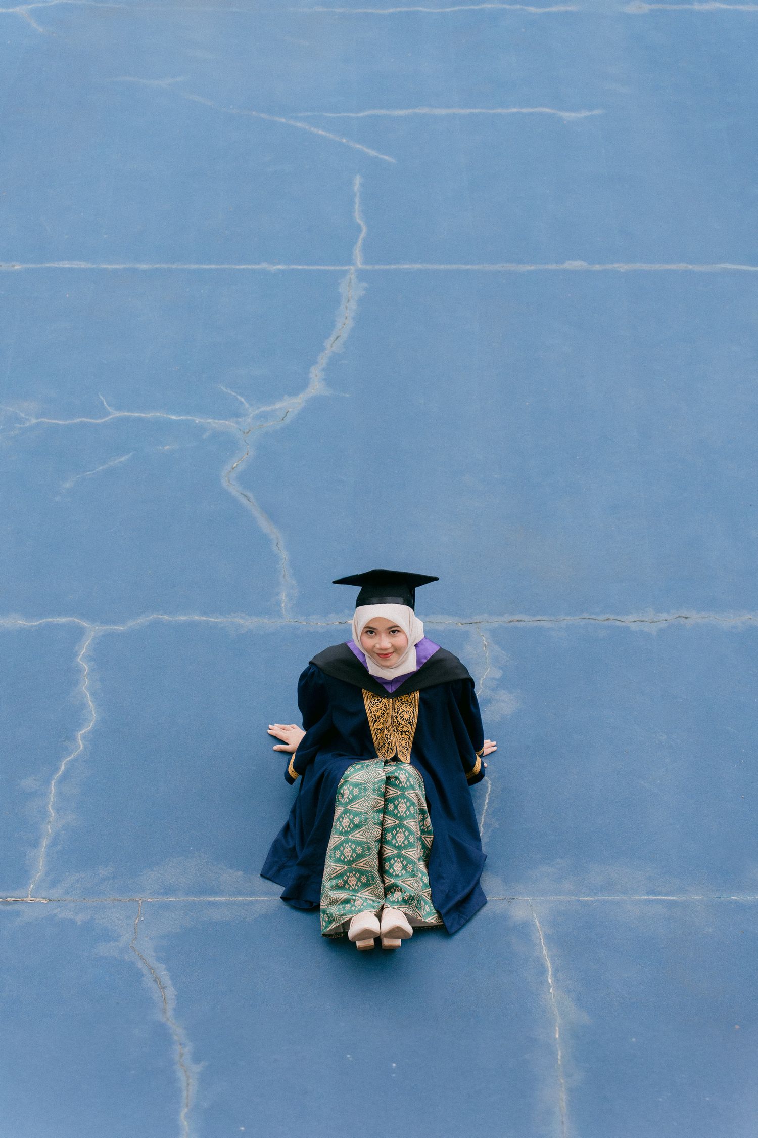 Aerial view of graduate in cap and gown lying on blue painted concrete surface creating minimalist composition.