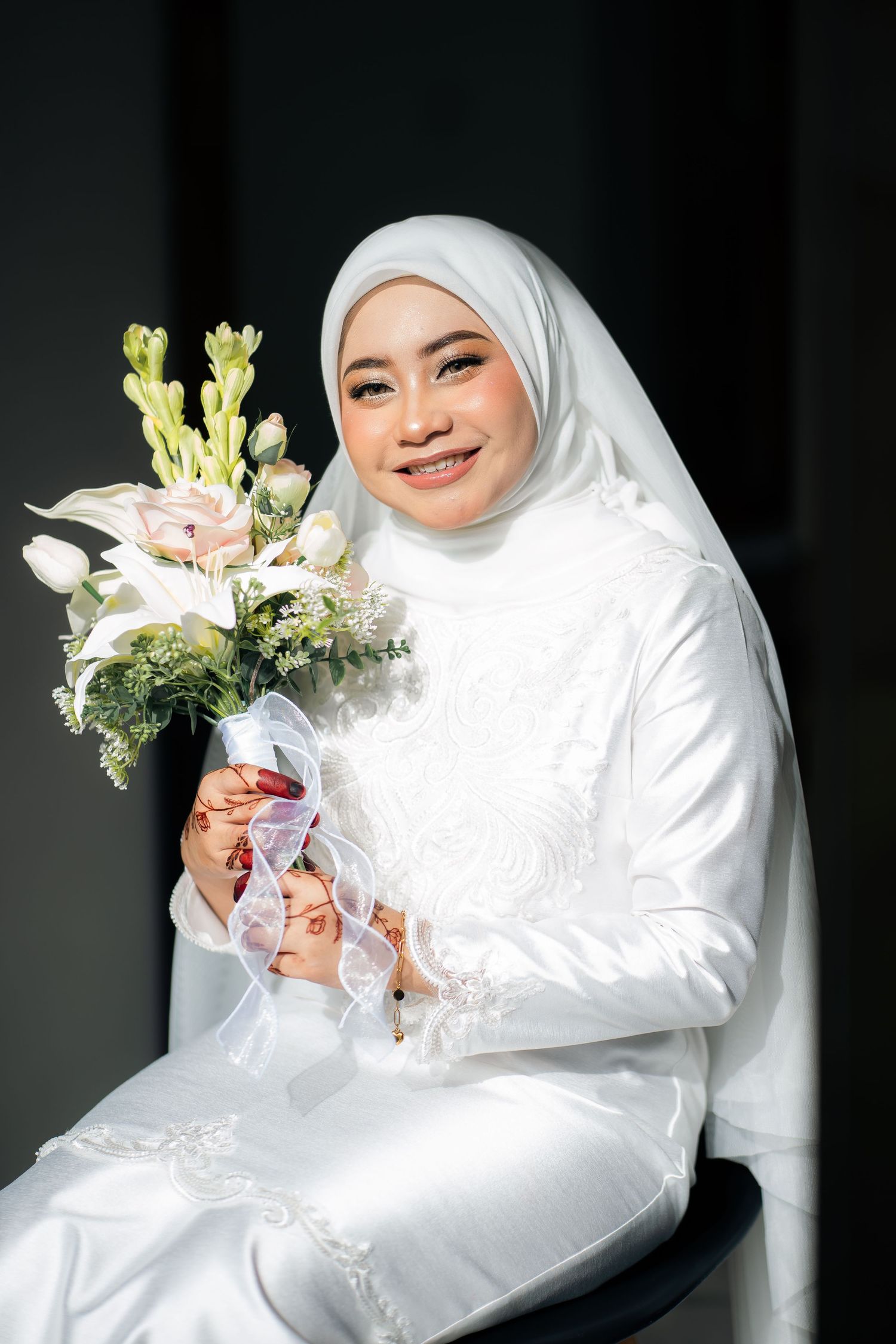 Elegant bride in white traditional dress and hijab holding a delicate bouquet of pink lilies and white flowers.