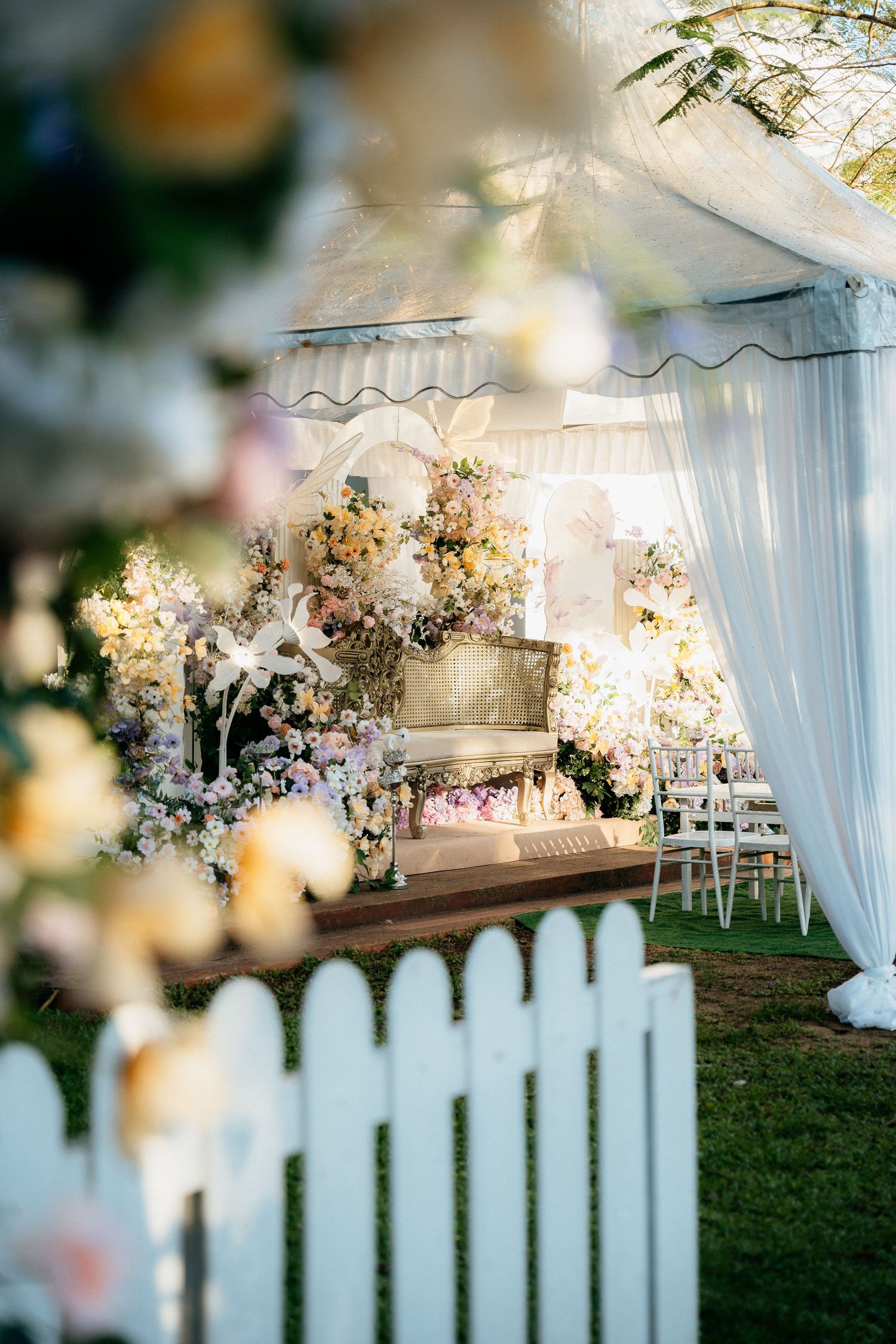 Elegant outdoor wedding reception setup with white draped tent, floral decorations, and white picket fence in foreground.