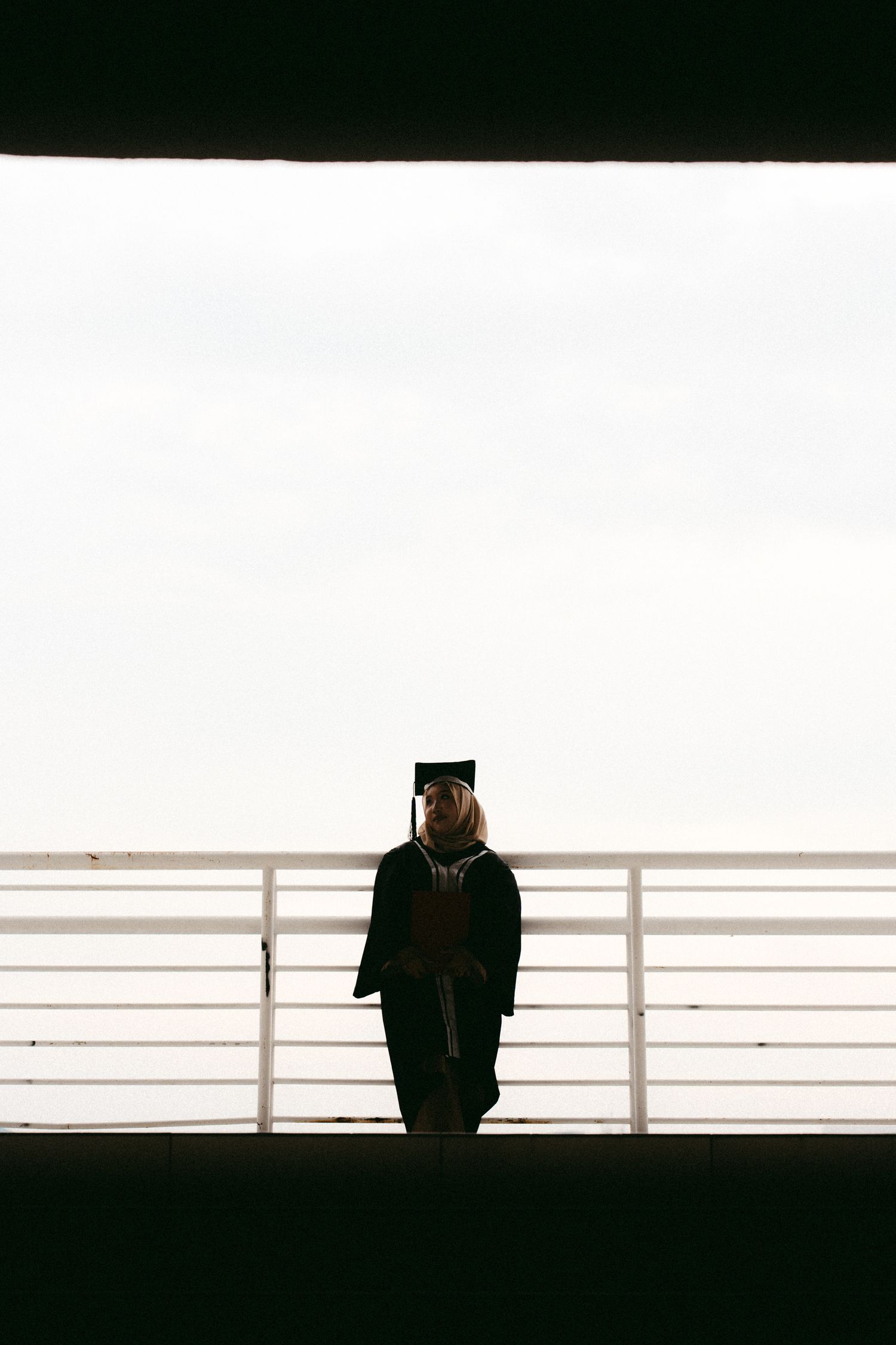 Graduate stands in silhouette against bright background wearing cap and gown with railing in foreground.