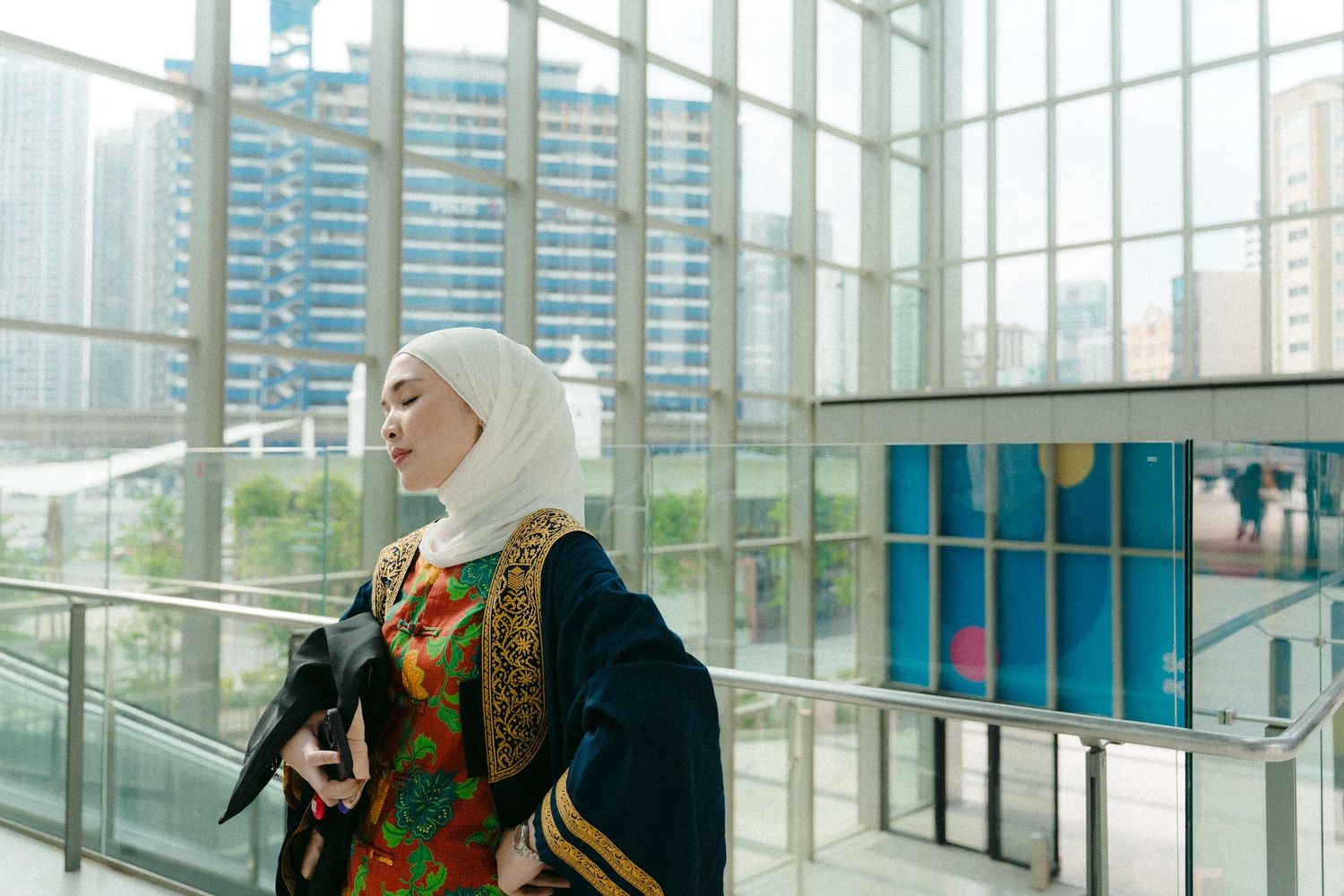 A graduate in traditional academic dress stands in a modern glass building with city views.