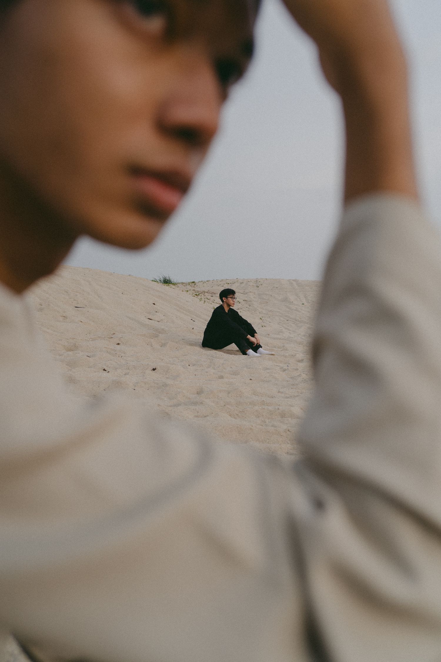 Artistic photo composition showing a figure sitting alone on sandy dunes through foreground framing.