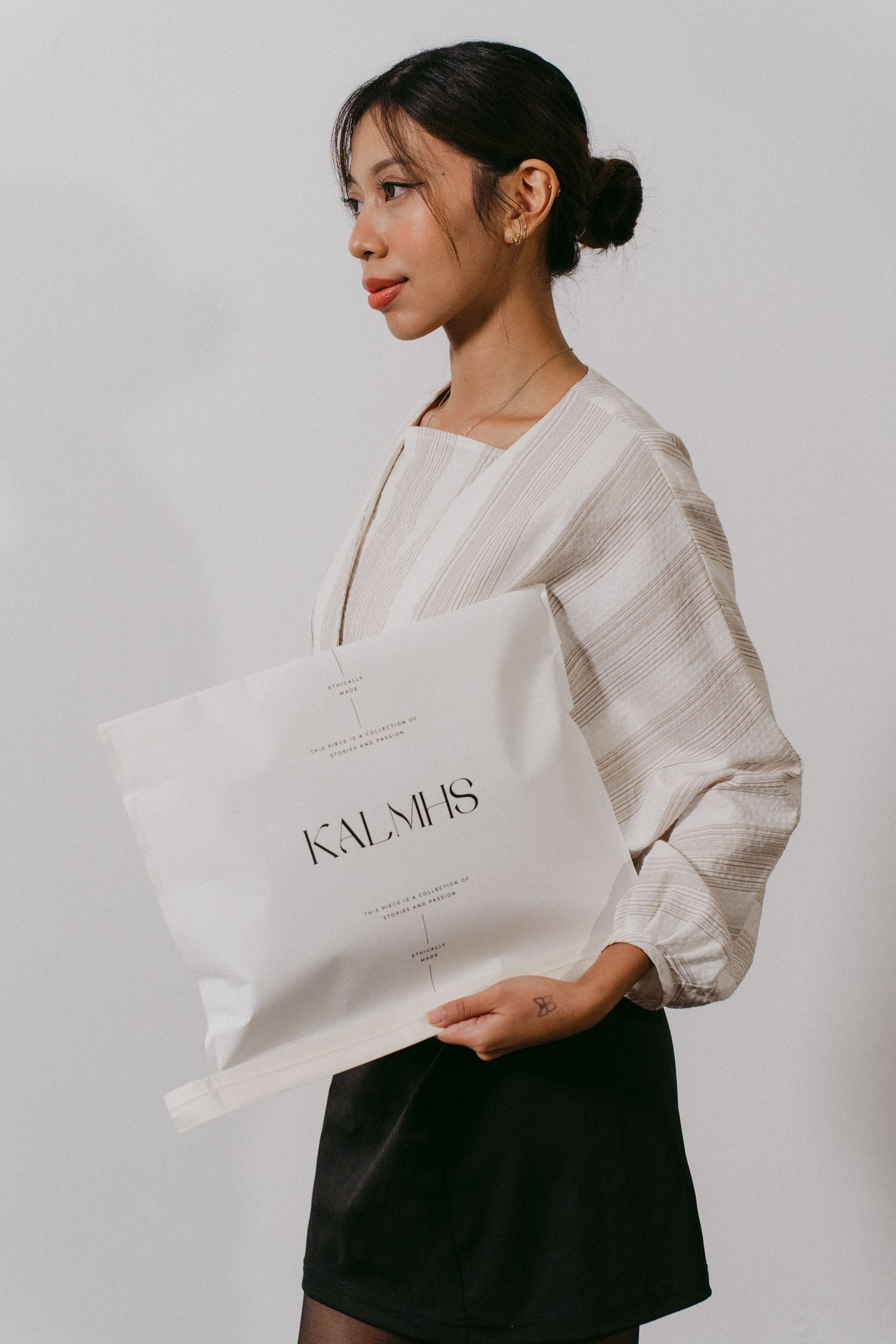 Fashion model wearing white blouse holding minimalist shopping bag against gray background.