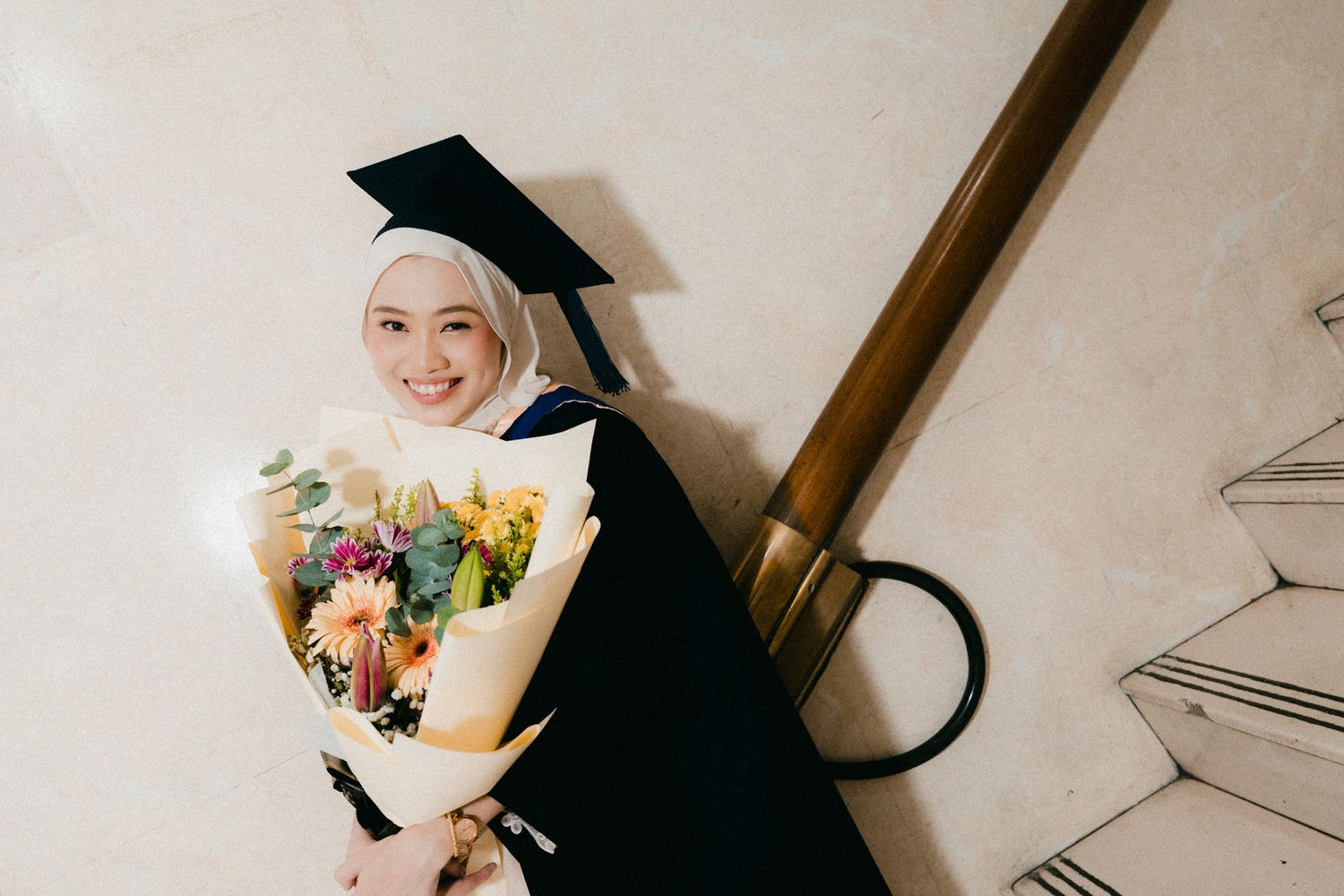 Graduate in black cap and gown holding colorful bouquet of flowers while sitting on white stairs.