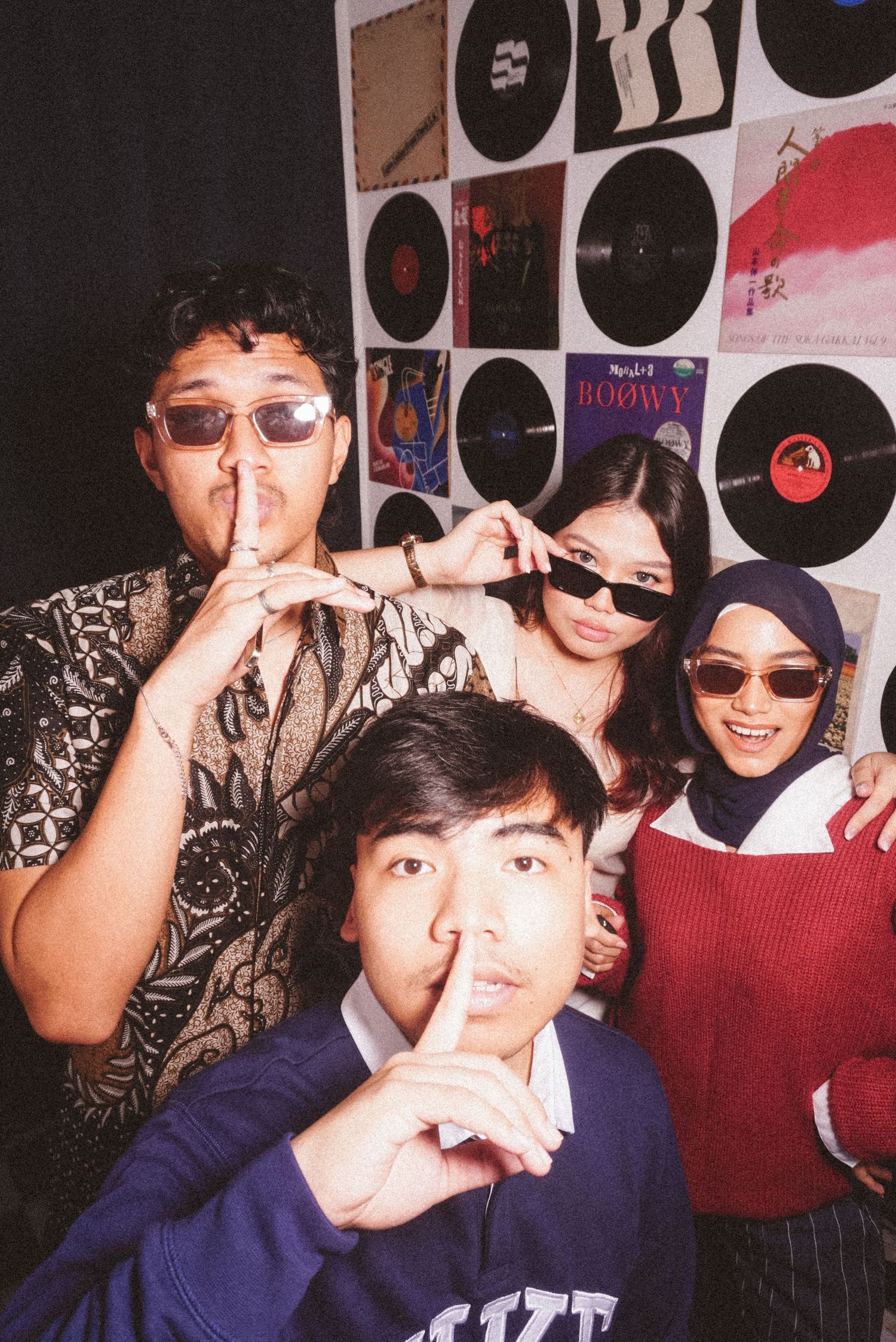 Four young friends pose playfully with fingers on lips in front of a vinyl record wall display.