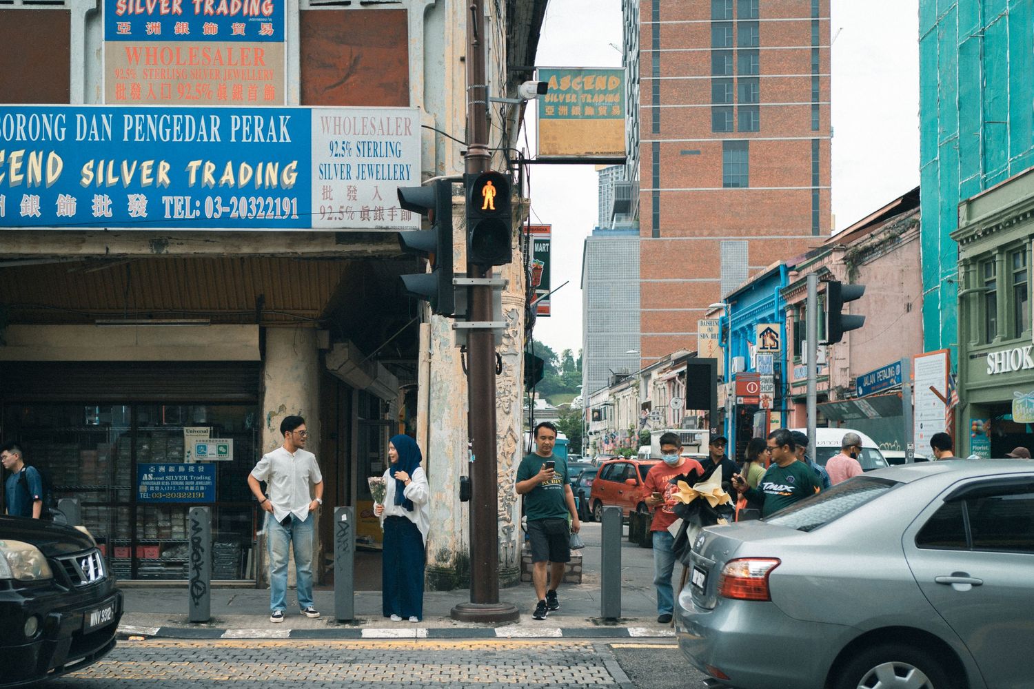 Two people wait at a street corner in front of businesses with Chinese signage.