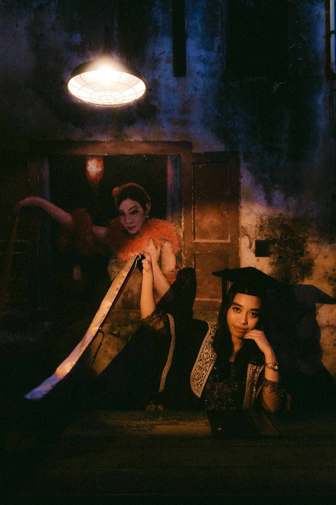 Two women in dramatic costumes pose in a dimly lit, atmospheric setting with a hanging lamp overhead.