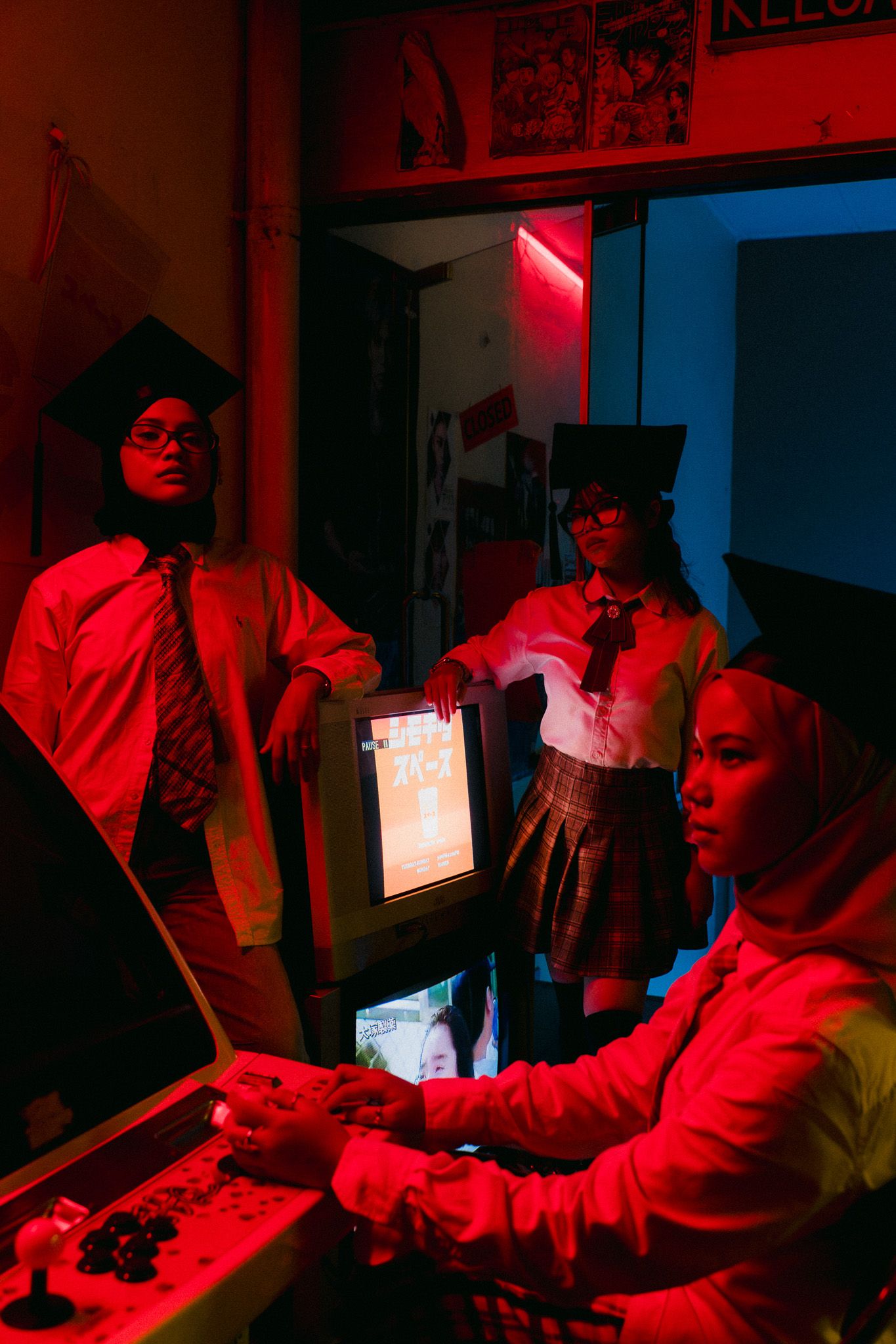 Three people in graduation caps pose around an arcade game cabinet under dramatic red neon lighting.