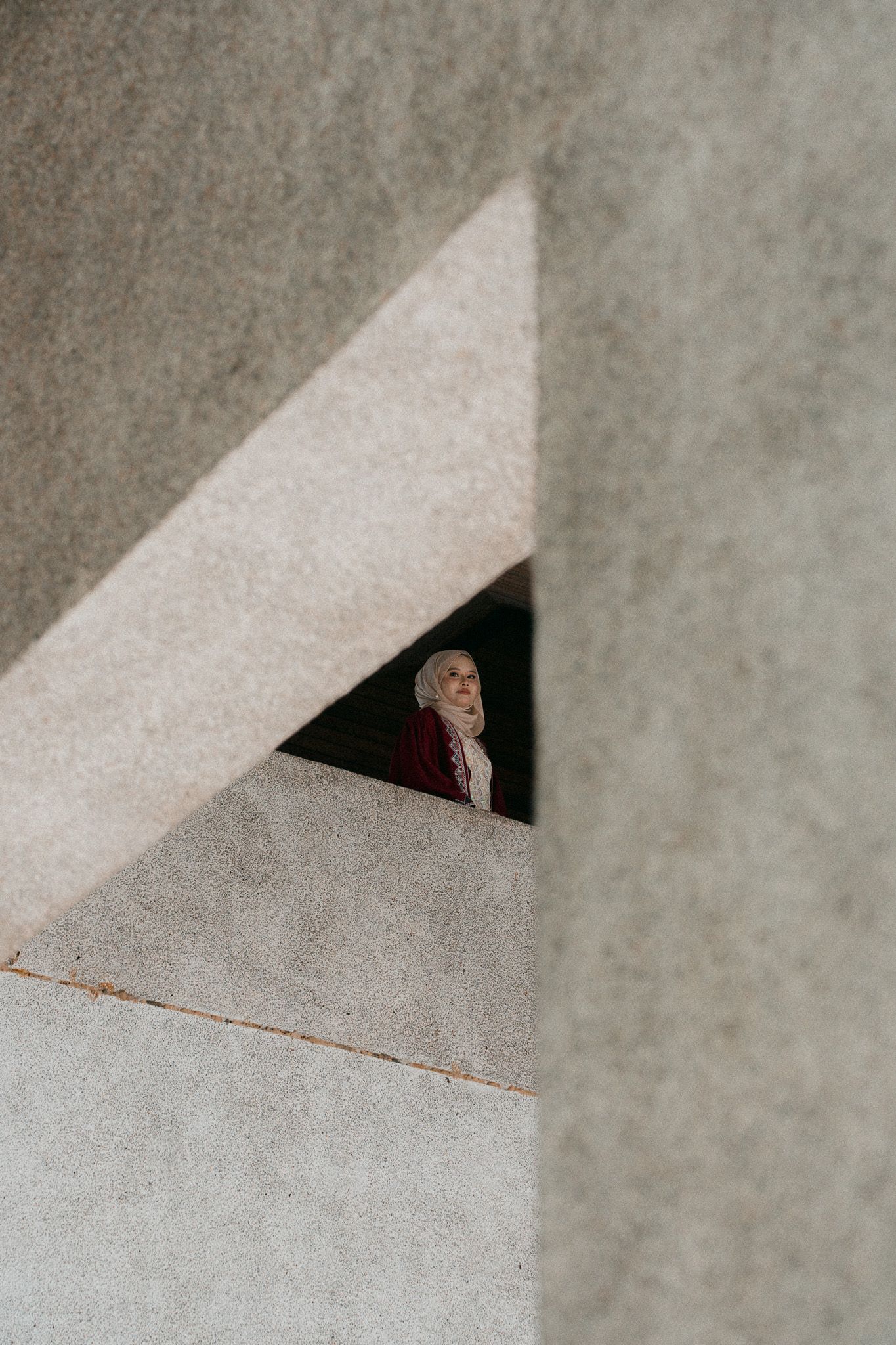 A woman wearing a hijab peers through a geometric concrete architectural opening in a minimalist structure.
