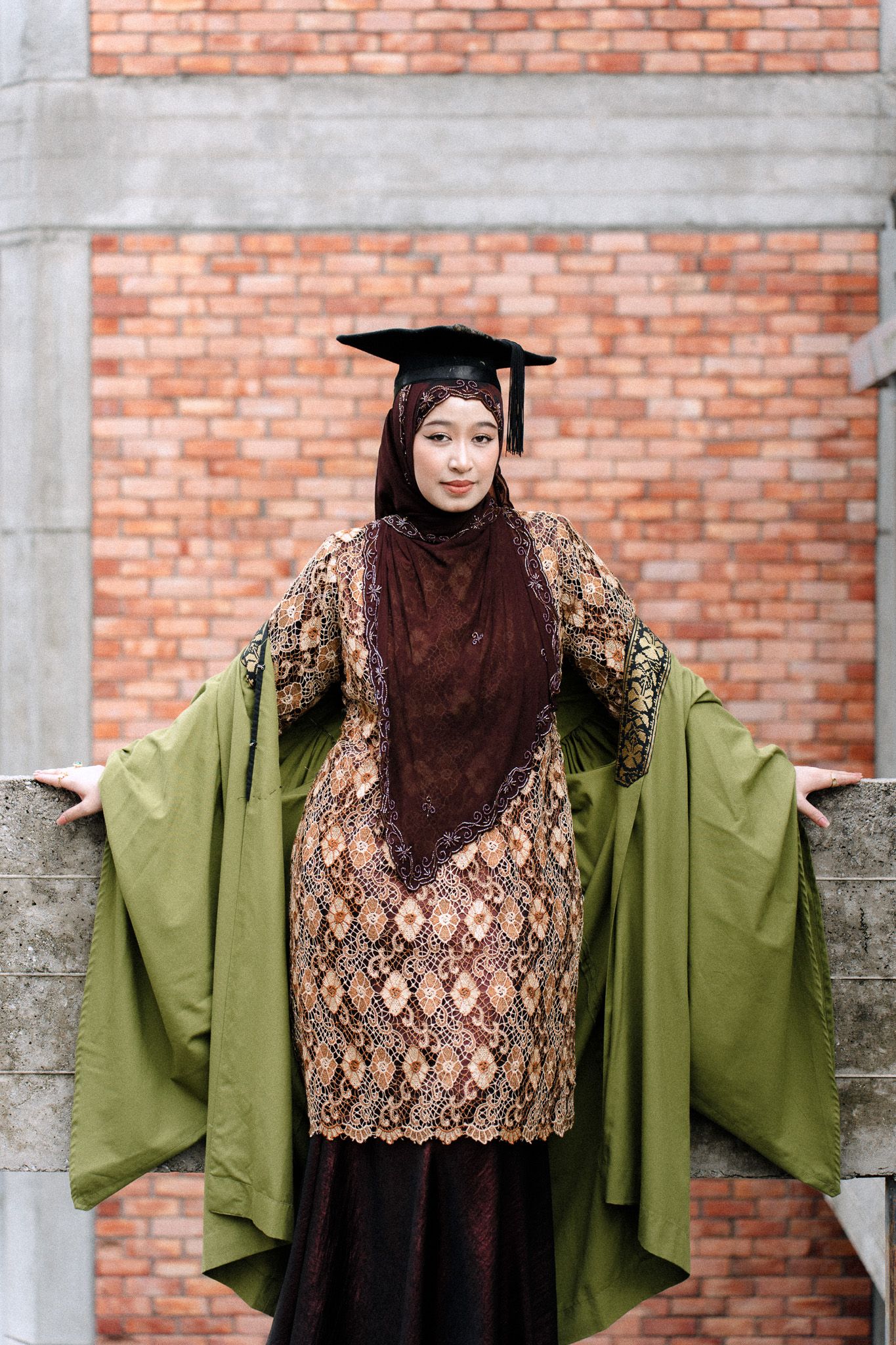 Muslim woman in hijab and graduation cap wearing traditional batik dress, posing proudly in front of brick wall.