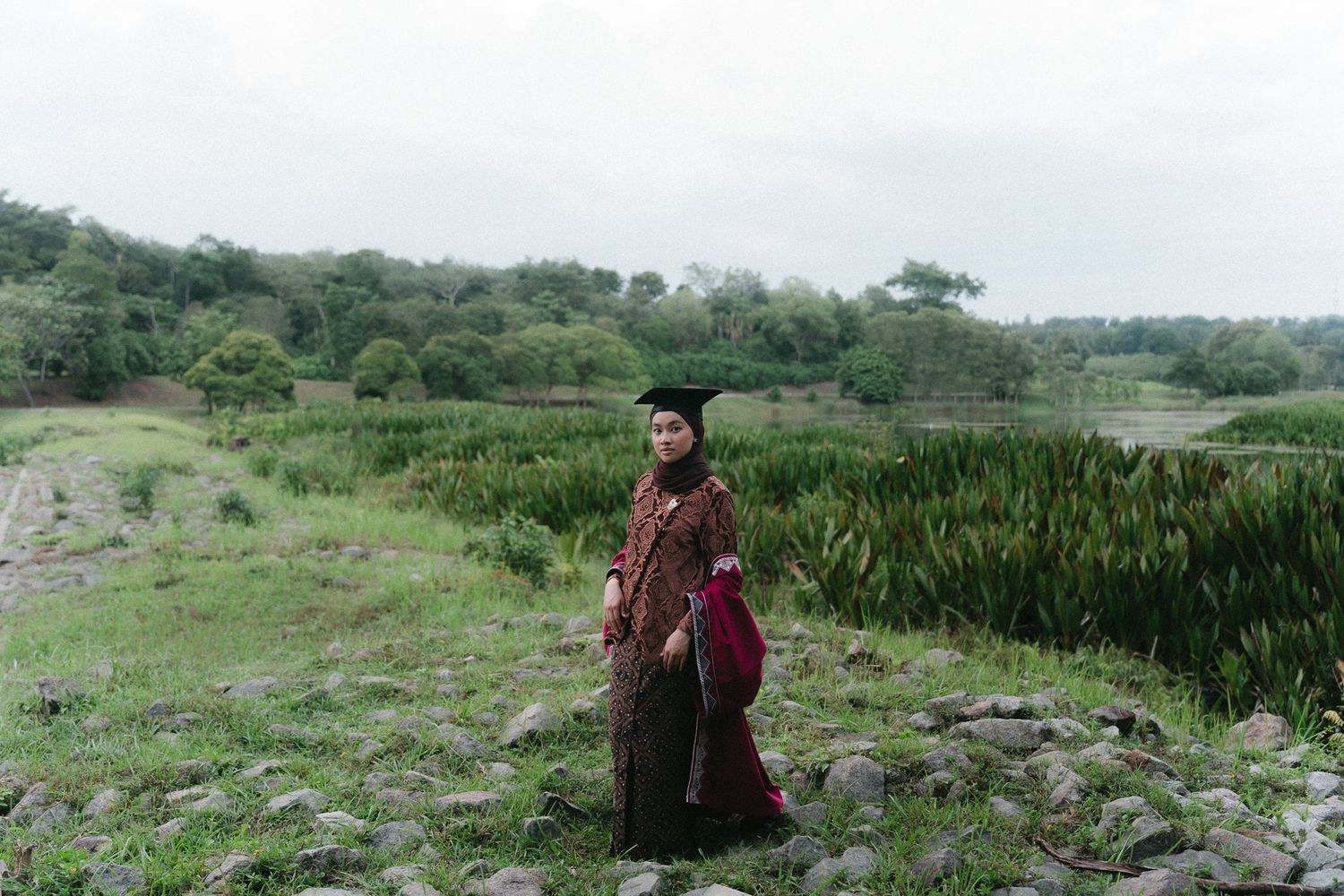 A person in traditional batik attire and graduation cap stands on rocky terrain with lush green landscape behind.
